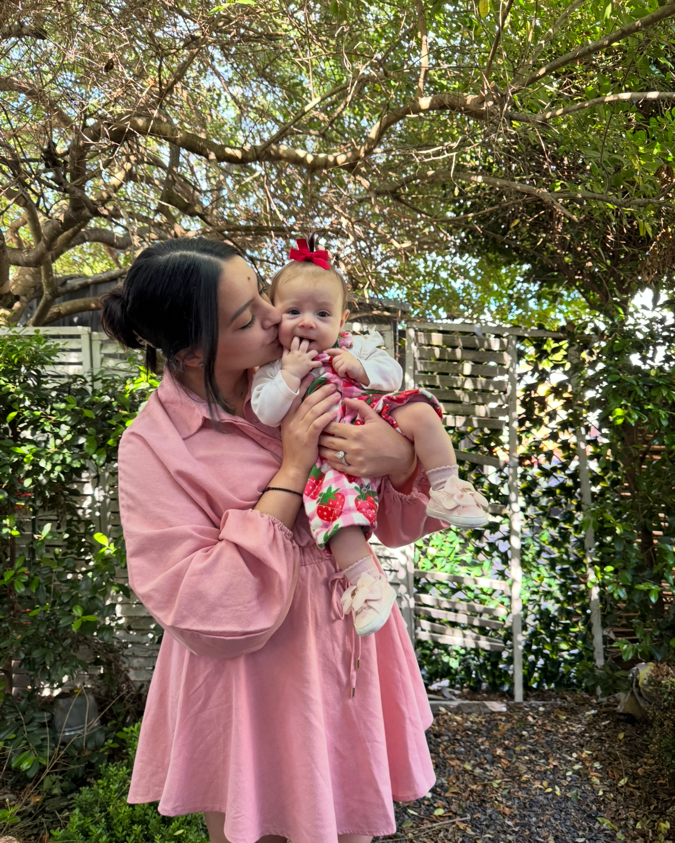 A young woman in a pink dress kissing a baby girl on the cheek outdoors, with trees and a wooden fence in the background.