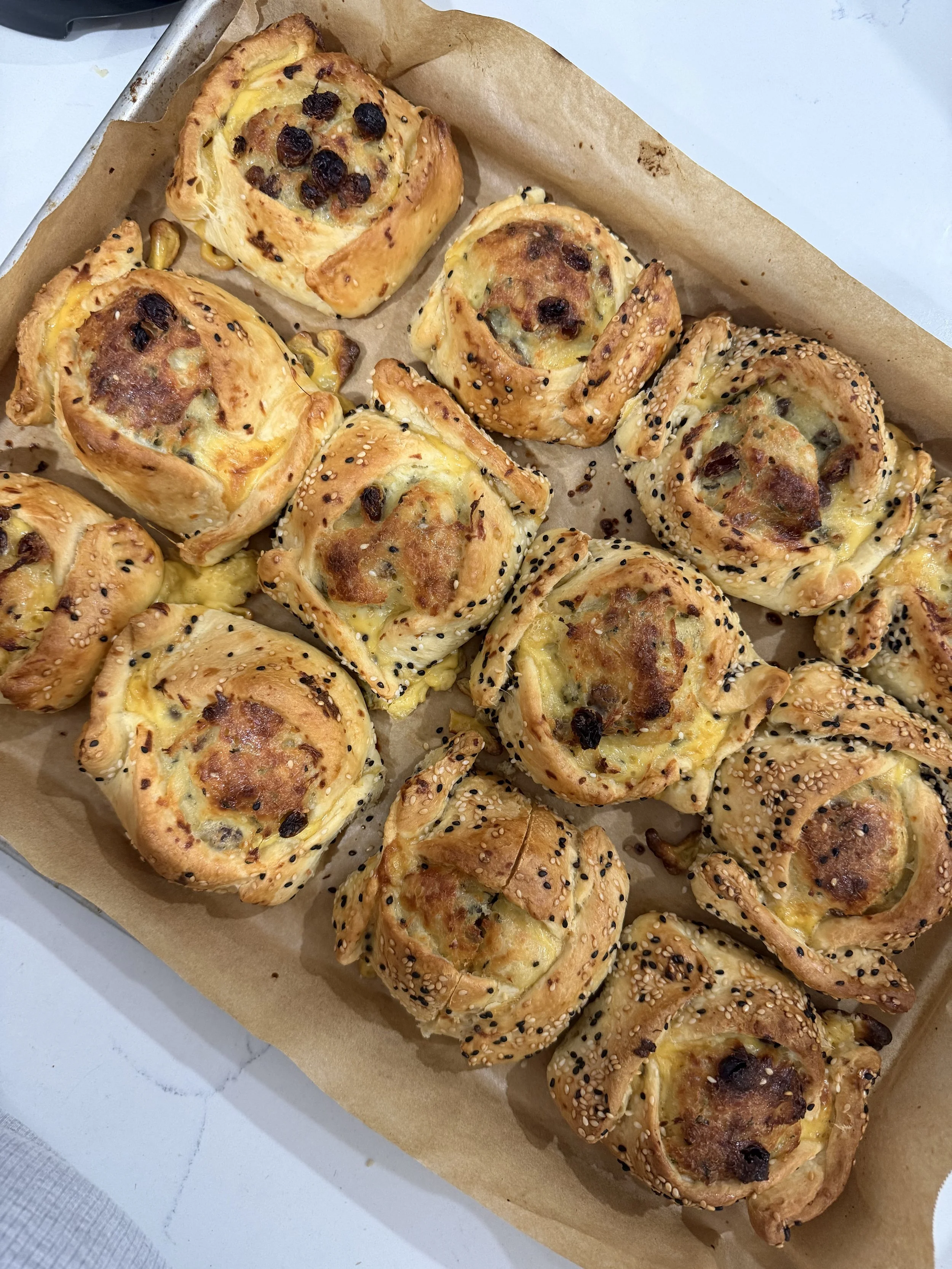 Baked savory pastries with diced vegetables, cheese, and black sesame seeds on top, arranged on parchment paper in a baking tray.