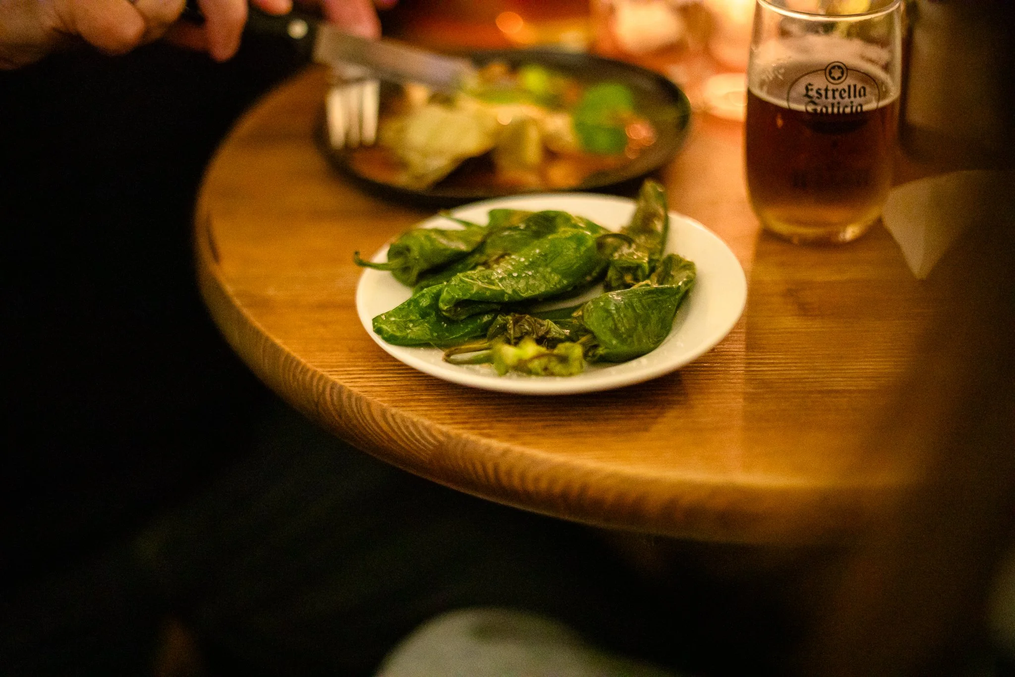 A white plate with cooked jalapeño peppers on a wooden table, with a person cutting food in the background, and a glass of dark beer nearby.