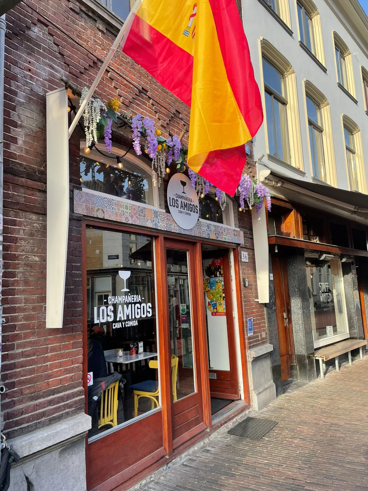 The exterior of a restaurant named 'Champanería Los Amigos' with brick walls, a Dutch door, and decorative purple flowers with string lights. A large Spanish flag hangs above.