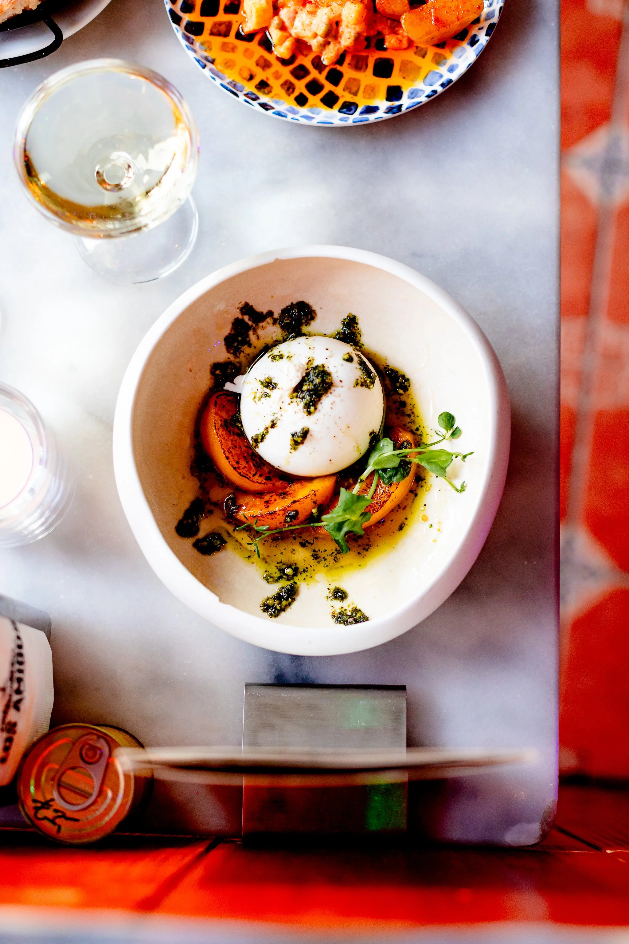 A bowl with sliced tomatoes, a soft-boiled egg, green herbs, and drizzle of olive oil on a white marble table, with a glass of white wine and a bowl of food in the background.