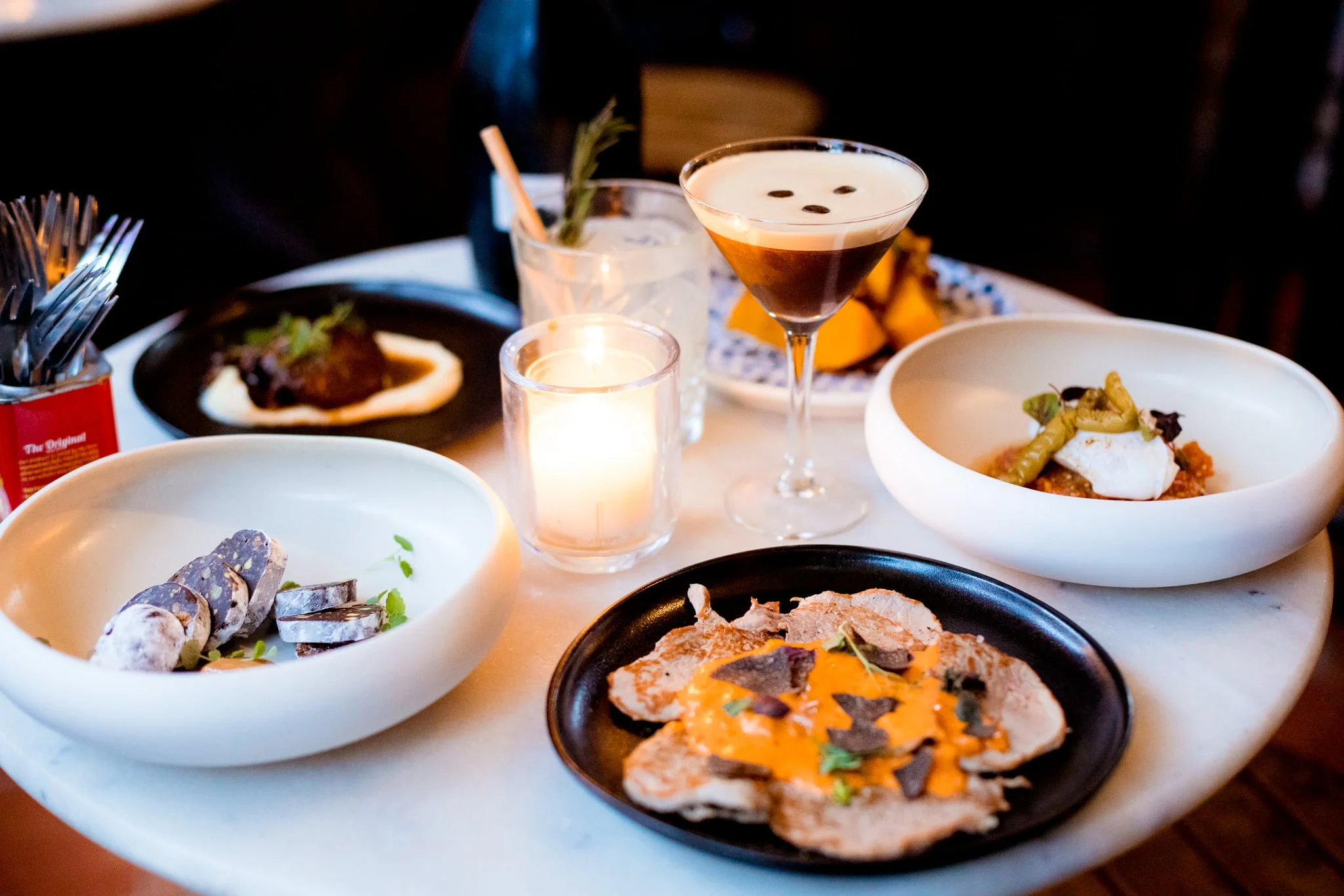 A table with various dishes, a candle, and a cocktail at a restaurant, featuring plates of meat, vegetables, and a decorated martini.