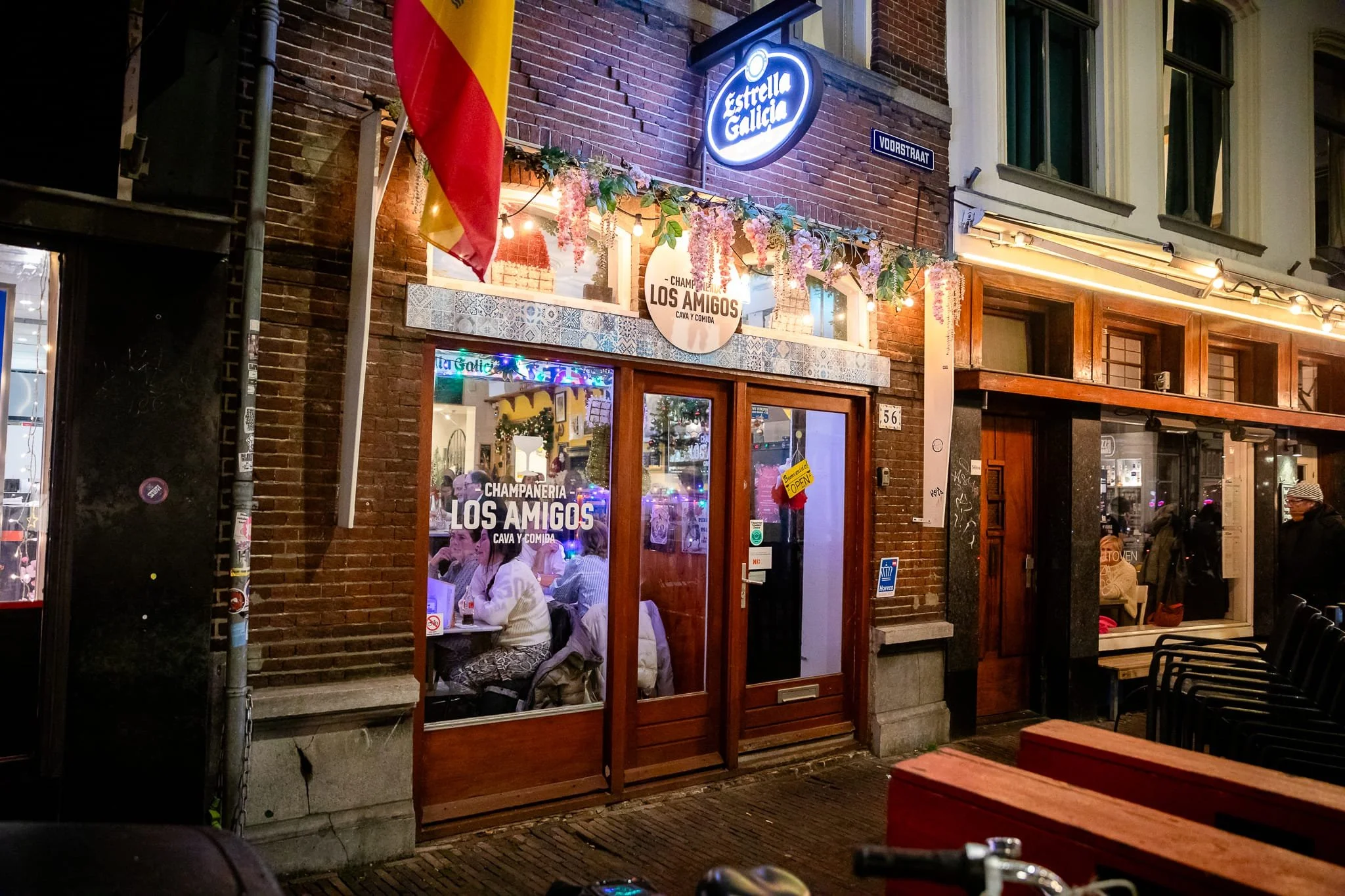 Nighttime view of a lively restaurant named 'Champaneria Los Amigos' on a busy street, with warm lighting, decorative flowers, and people dining inside, seen through large glass windows.