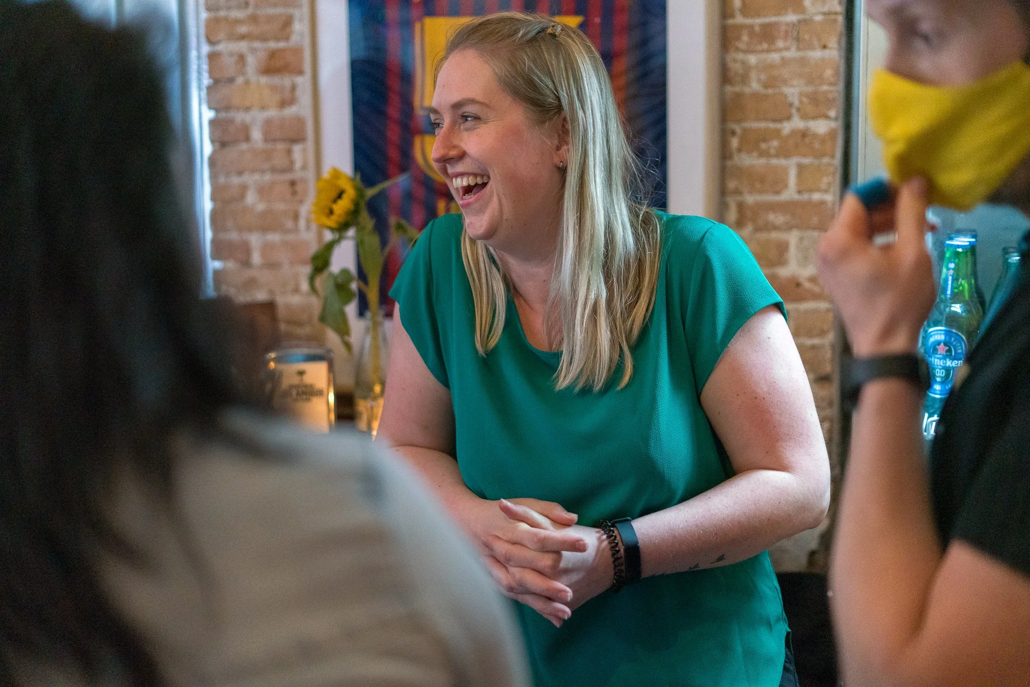 A woman with blonde hair, wearing a teal top, is smiling and engaging in conversation with others around her in a cozy cafe with brick walls and sunflower decor in the background.