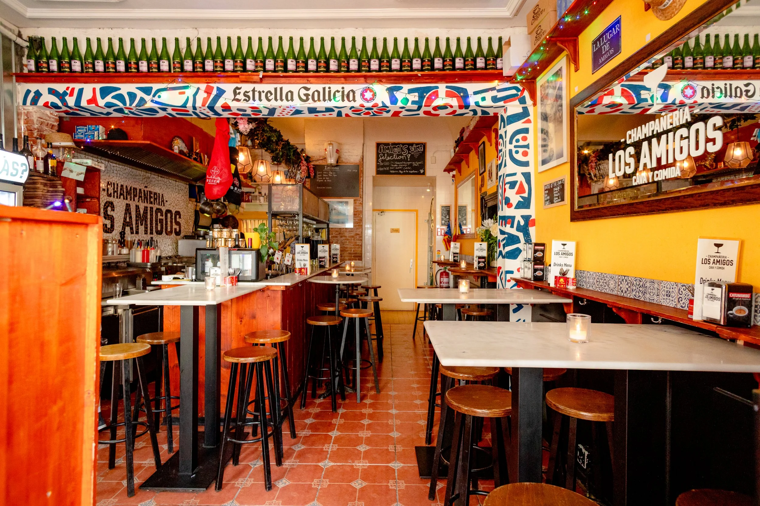 Interior of a cozy restaurant or bar with yellow walls, wooden and marble tables, bar stools, shelves with bottles, colorful decorations, signs, and a mirror; candlelit ambiance.