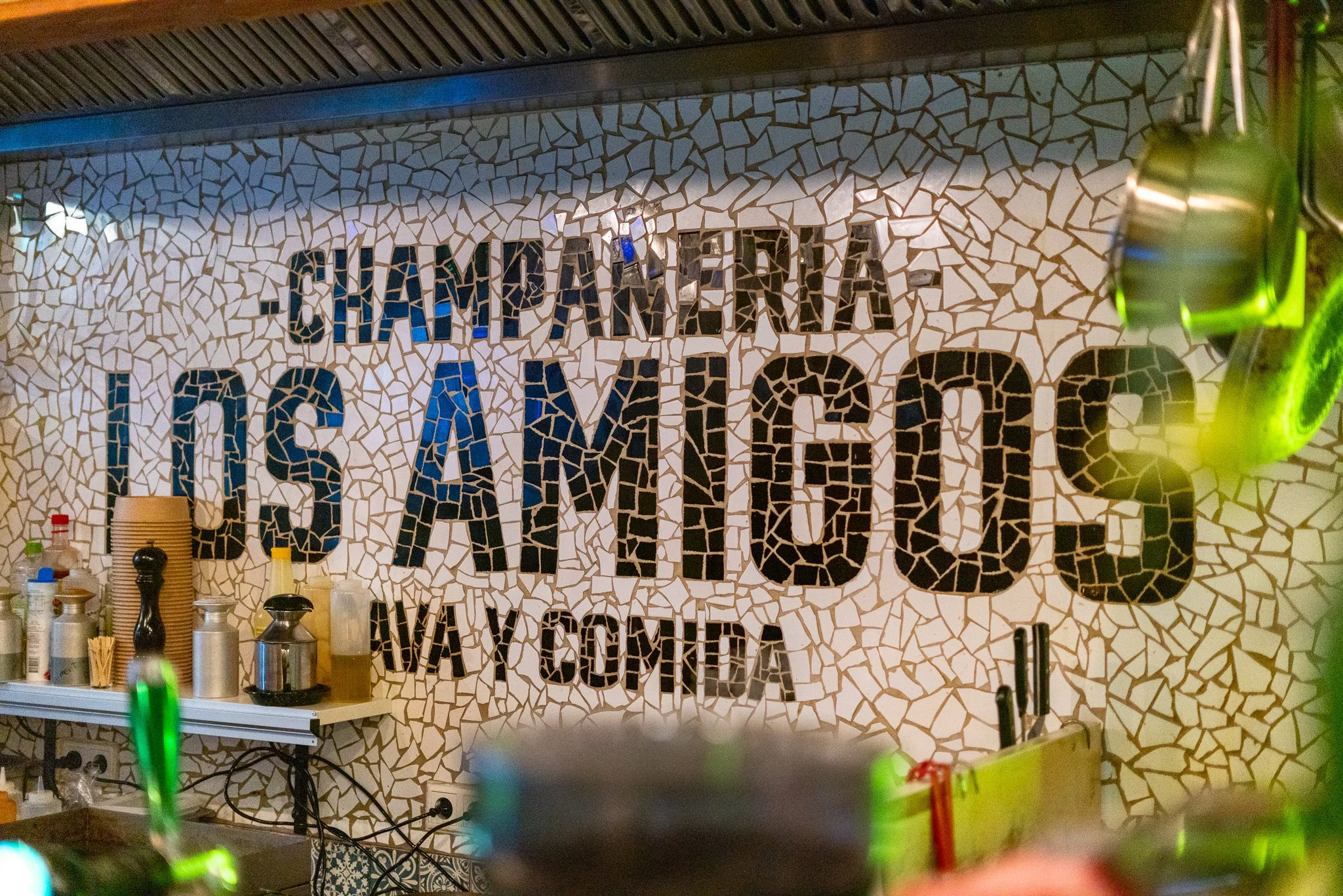 Mosaic wall with the words "Champingera Los Angeles" in large, black and blue letters, with smaller text "VIA COFTA" underneath, in a restaurant or cafe setting.