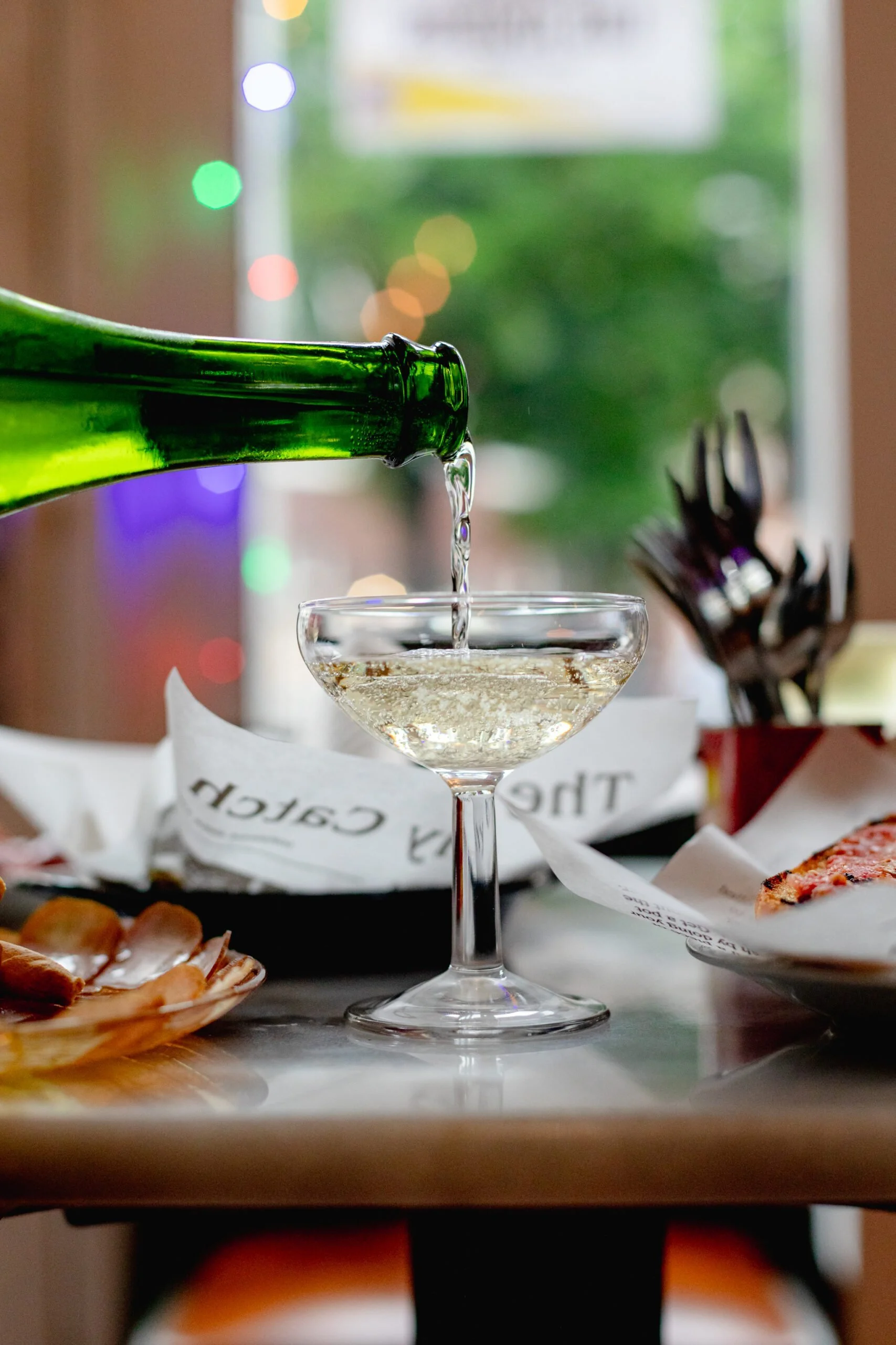 A glass of white wine being poured from a green bottle in a cozy restaurant setting with blurred colorful lights and dishes on the table.