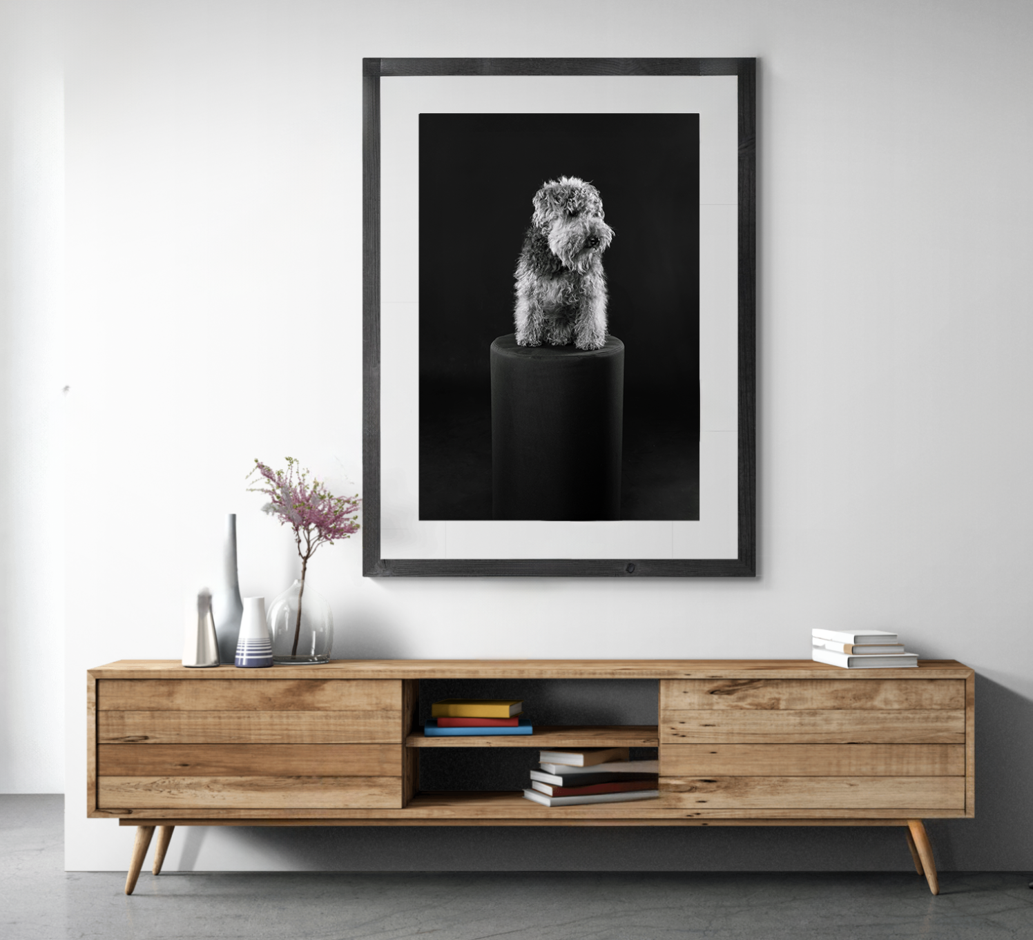 A black-and-white photograph of a fluffy dog sitting on a pedestal, displayed in a black frame on a white wall, with a wooden console table and vases with flowers in front.