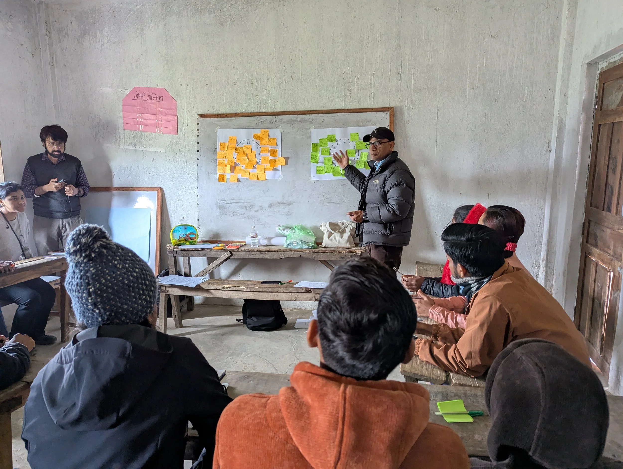 Participatory co-design session in a village classroom in the Terai region of Nepal, with a facilitator presenting sticky note timelines on the wall during field research on gendered nutrition norms by Lagori Collective with USAID