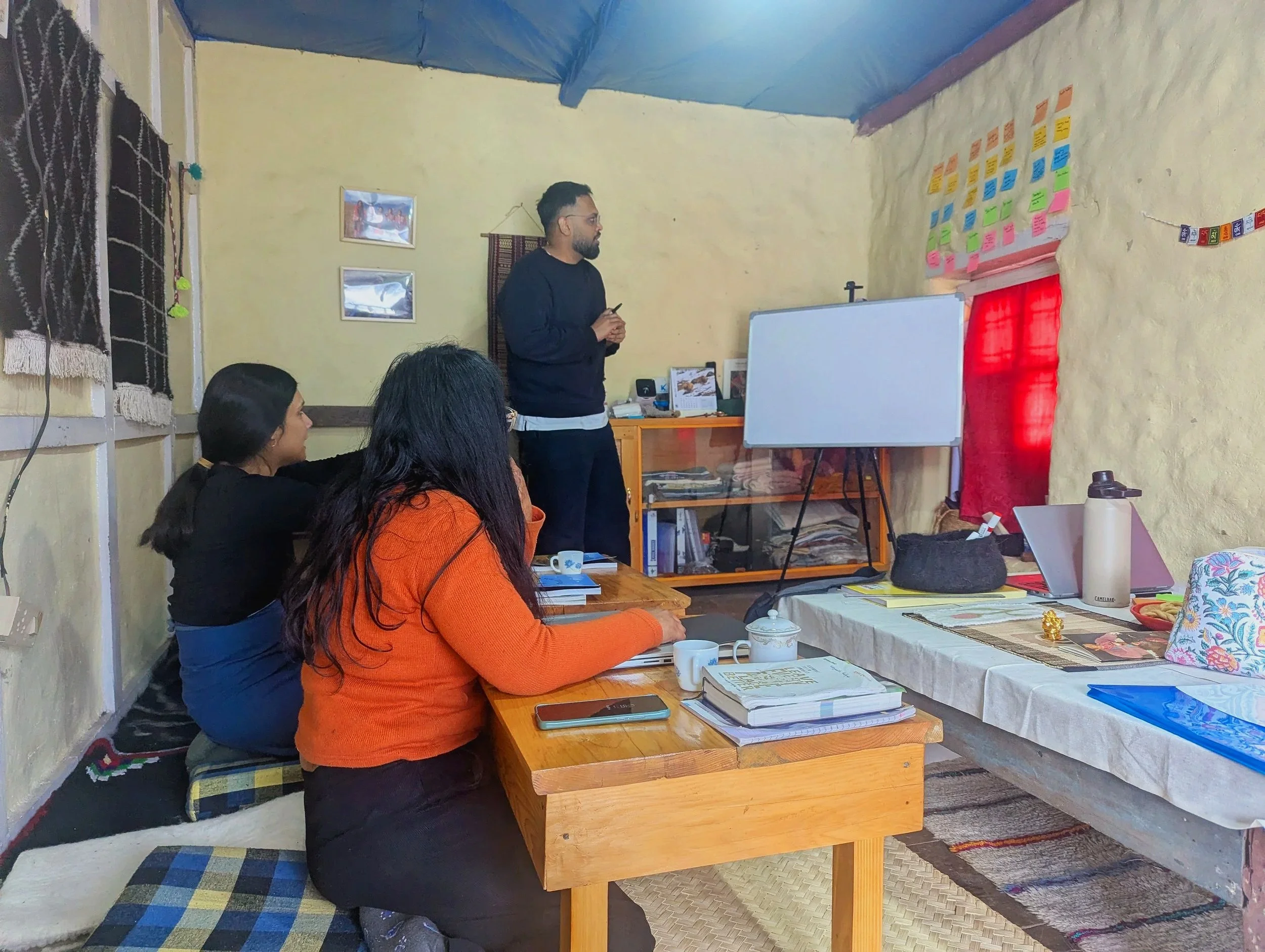 A man is standing and giving a presentation or lecture in a room while three women sit at a table listening. The room has a whiteboard, colorful sticky notes on the wall, and various papers and objects on the table and shelves.