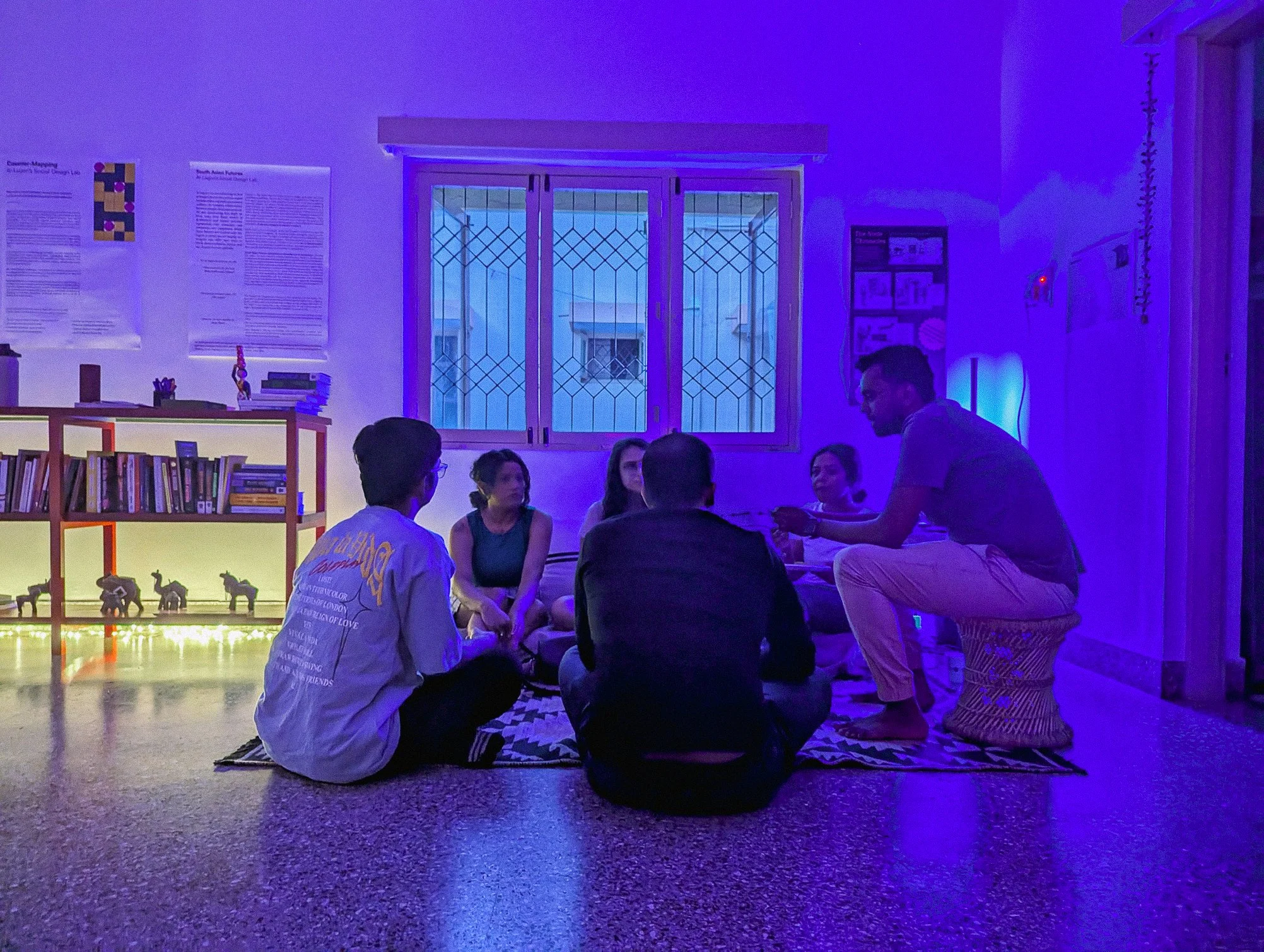 A small group seated in conversation on the floor of Lagori Collective's community space in Bengaluru during an evening gathering, lit in deep purple with bookshelves and exhibition material visible in the background.