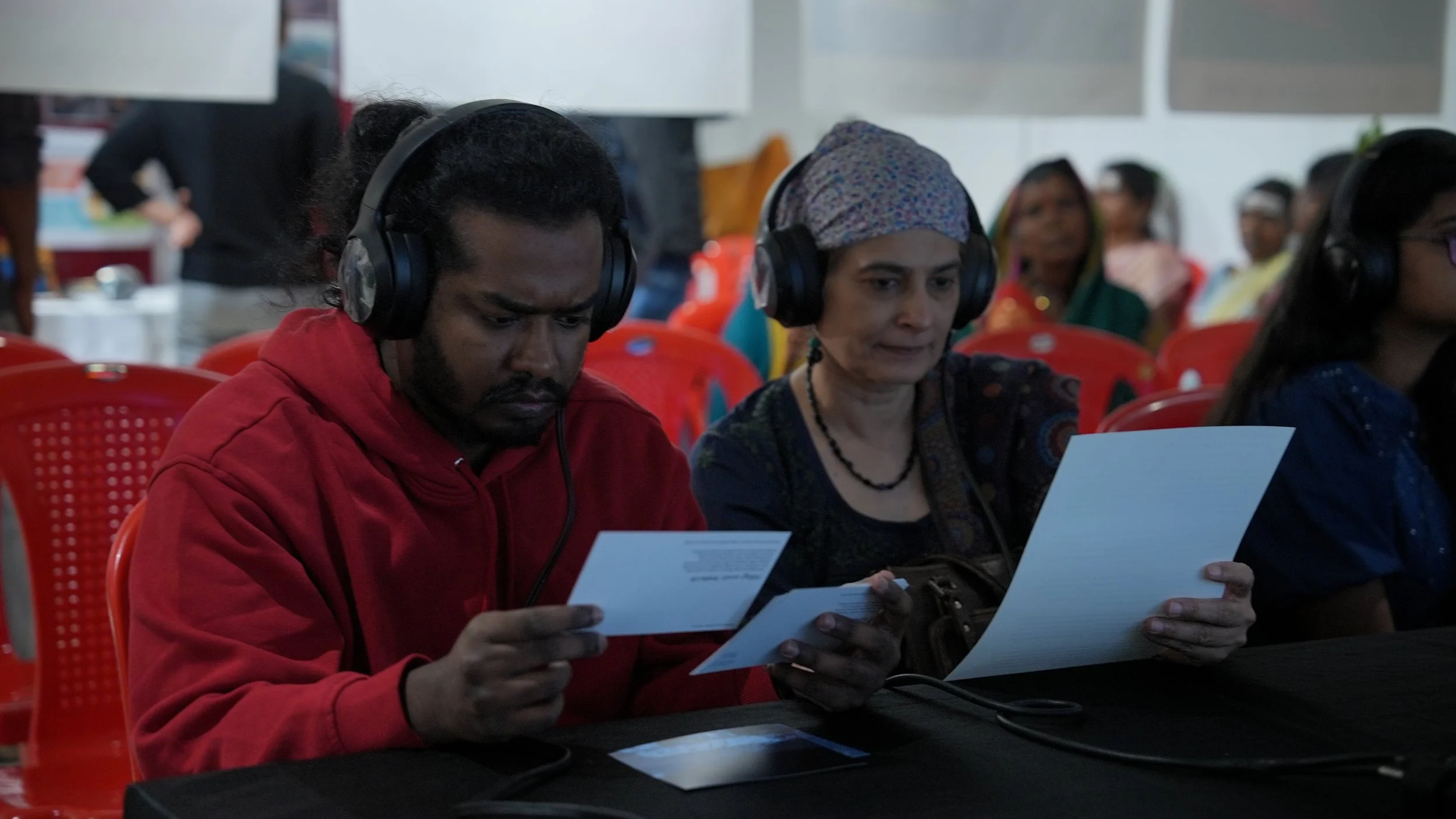 Two visitors wearing headphones reading accompanying text during the Words for Other Worlds installation by Lagori Collective, exploring South Asian ecological language and climate knowledge.