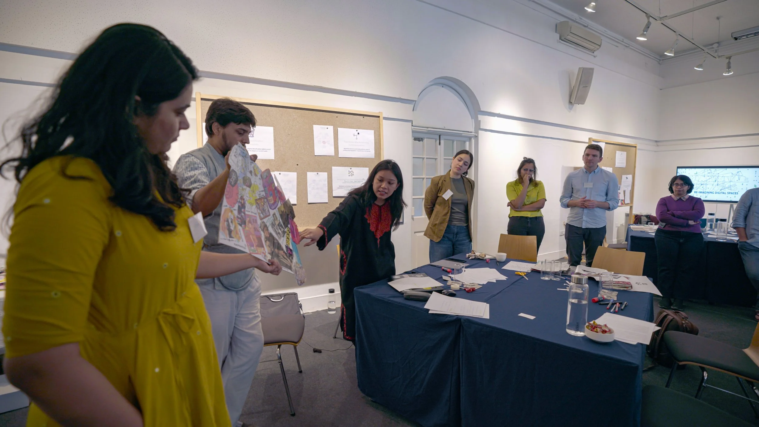 Group of seven people standing around a table with papers, markers, and glasses in a room with white walls and a bulletin board.