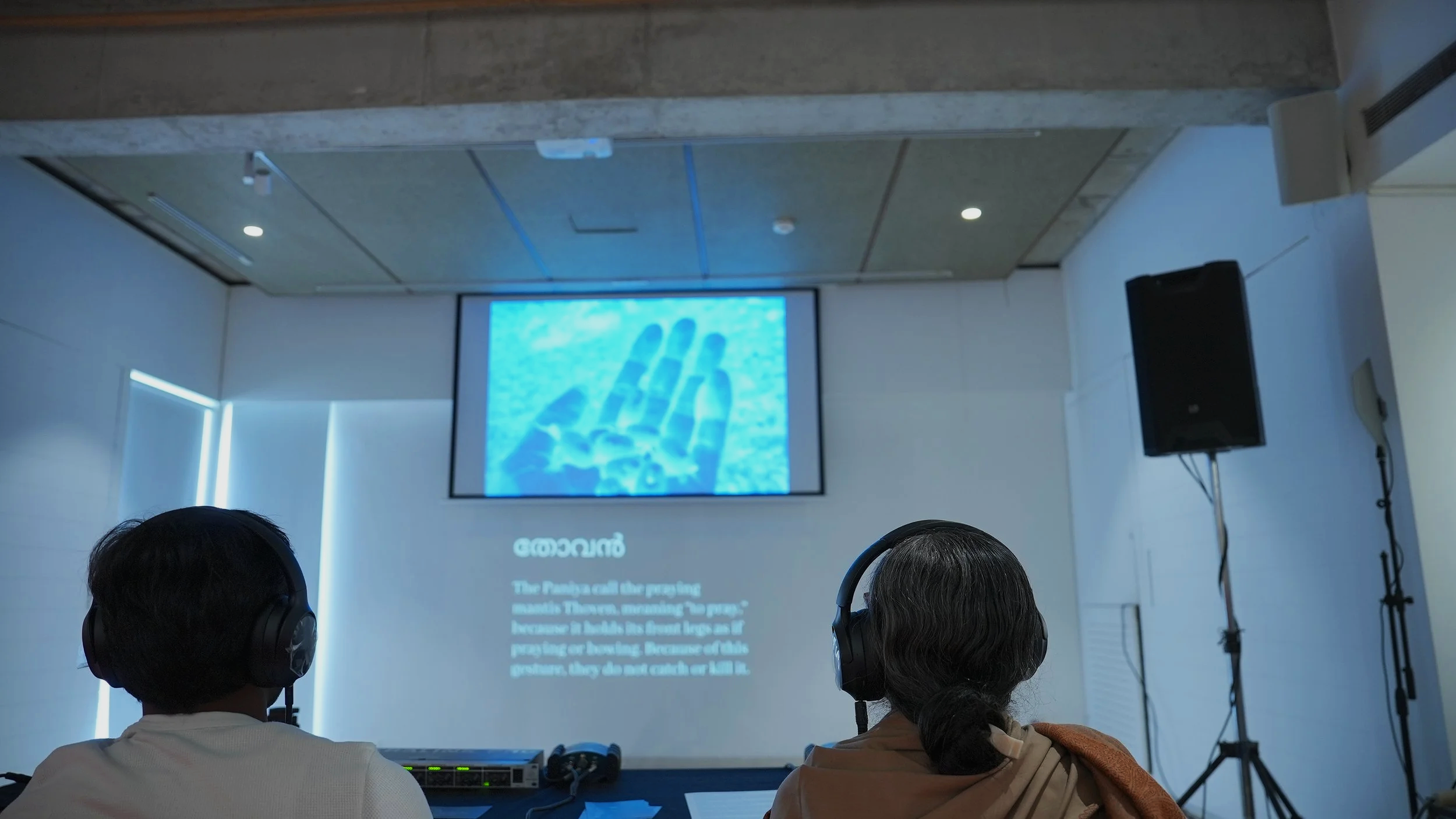 Two visitors wearing headphones watching a projected screen displaying Malayalam text and a close-up of a praying mantis during the Words for Other Worlds installation by Lagori Collective, TDU.