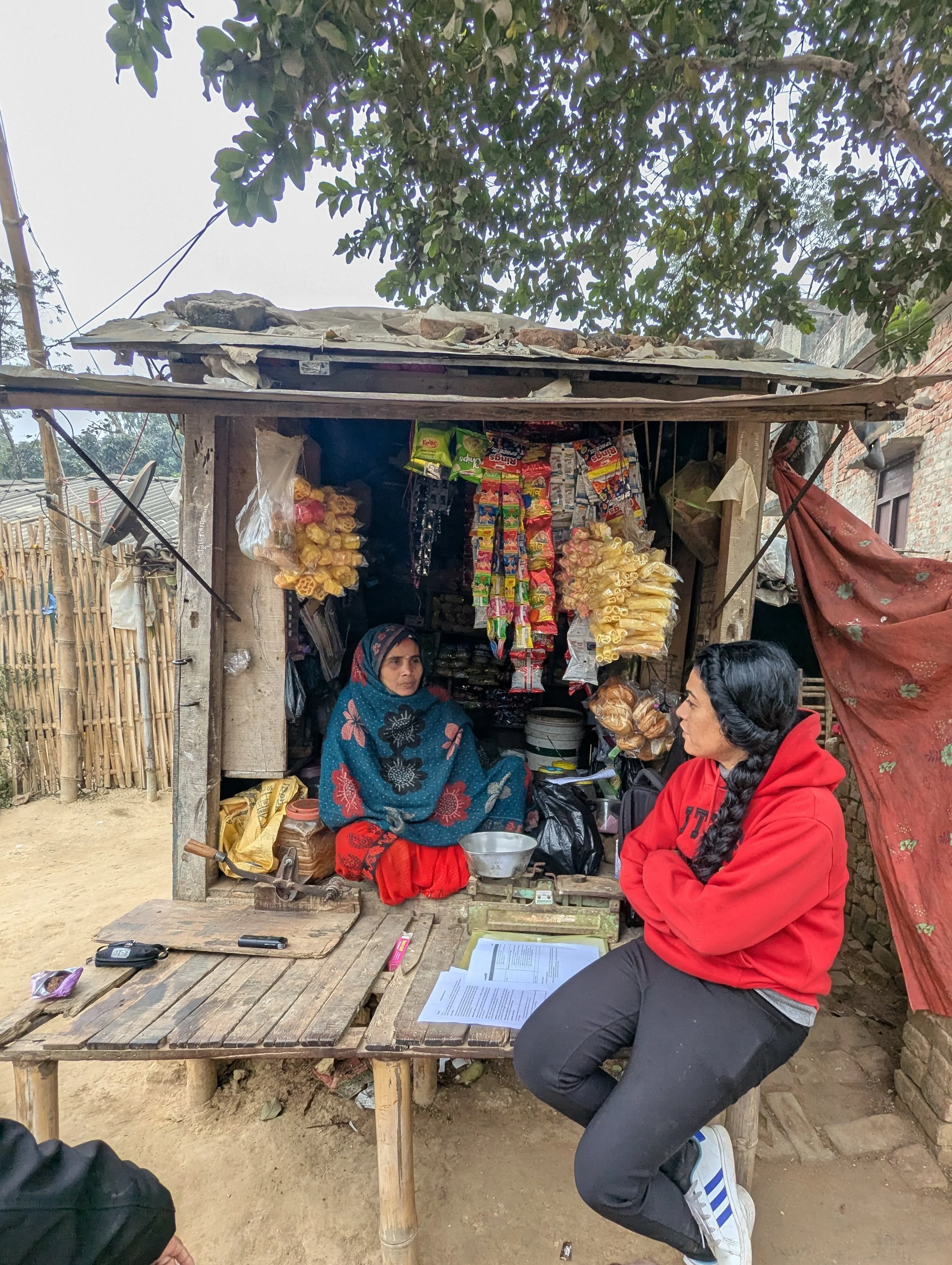 A researcher conducting a field interview with a woman shopkeeper at a roadside stall in rural Nepal, during field research on nutrition and behaviour change for the USAID Integrated Nutrition project by Lagori Collective, Helen Keller International.