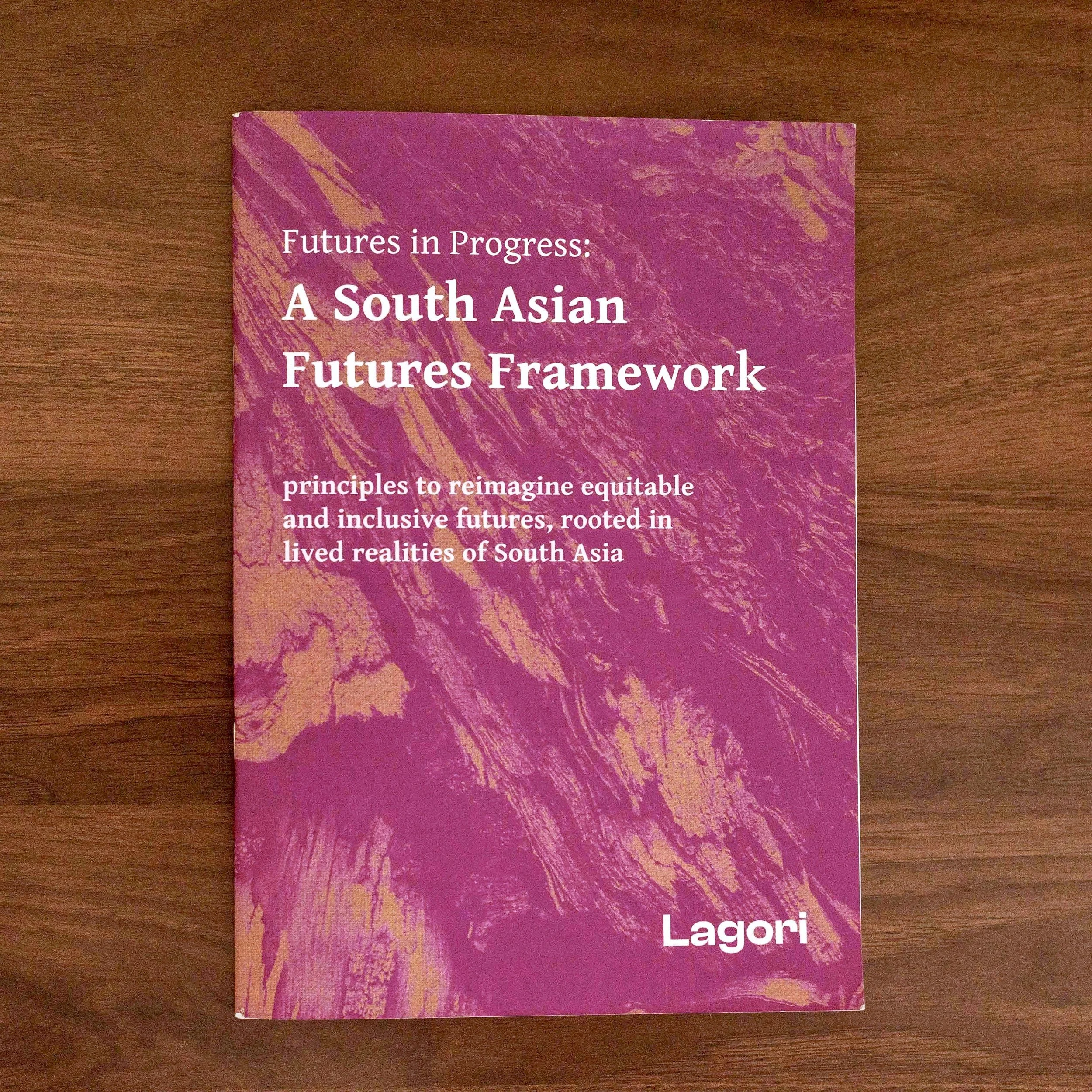 Physical copy of Futures in Progress: A South Asian Futures Framework, a paperback publication by Lagori Collective, photographed on a wooden surface showing the purple and gold textured cover.