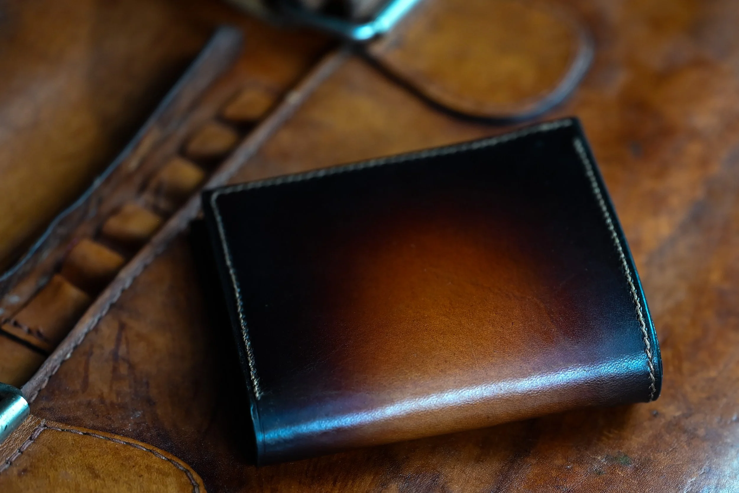 Portefeuille en cuir de la collection Héritage, fermé, patine dégradée du brun au noir visible sur la couverture, posé sur une sacoche en cuir ancienne des années 1950.