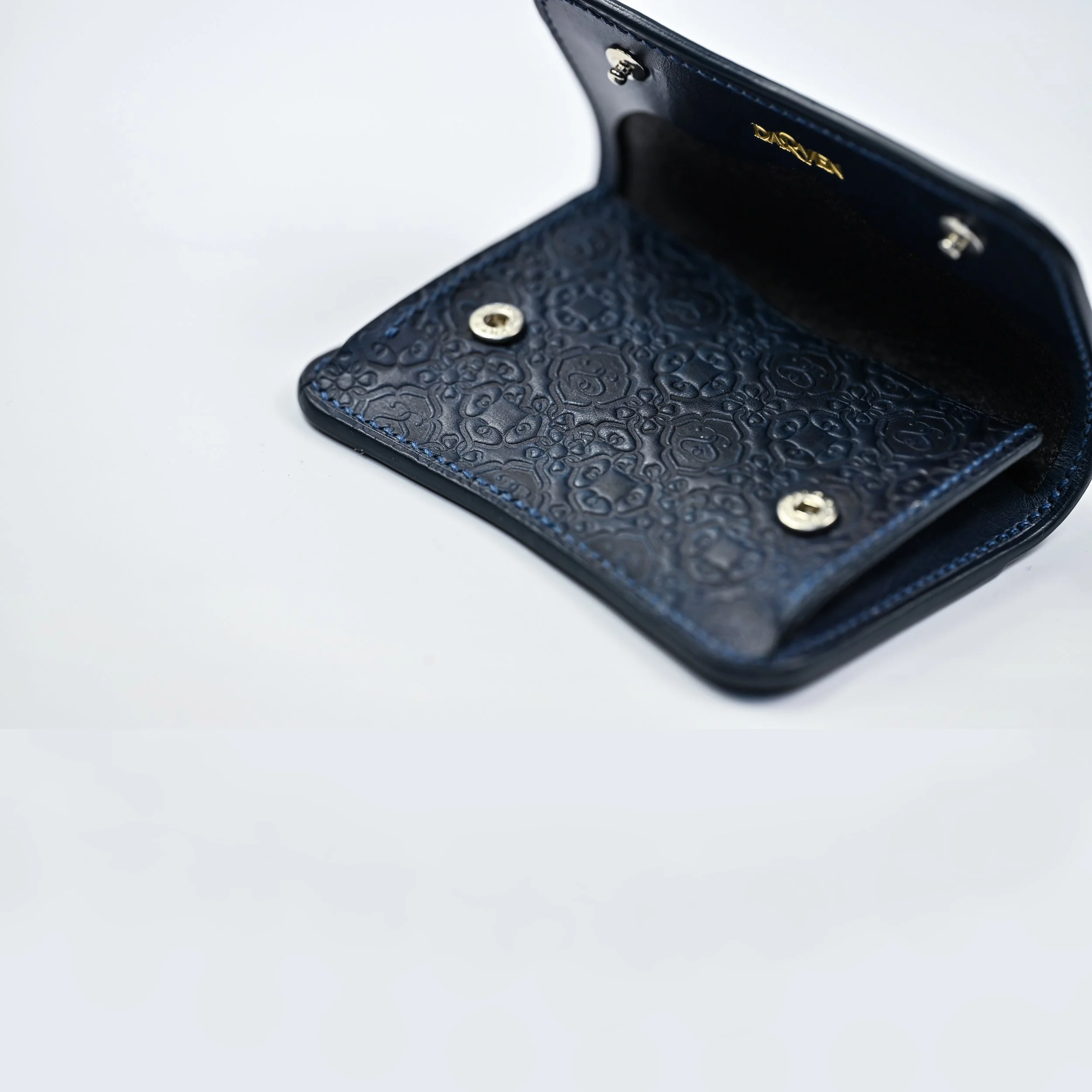 Une petite pochette en cuir bleu ouverte, montrant l'intérieur avec un motif en relief, sur un fond blanc.