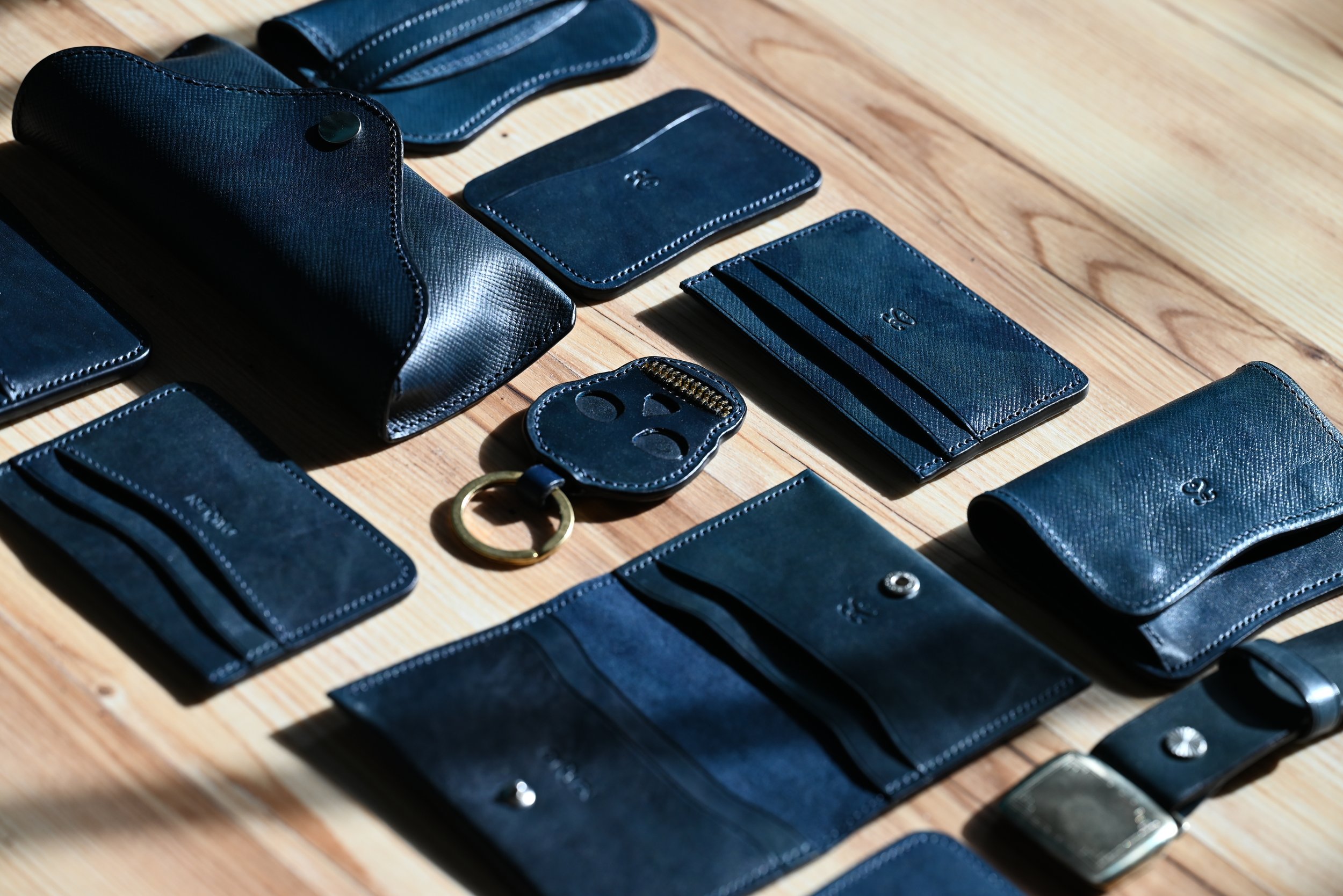 Ensemble d'articles de la collection Indigo en cuir teinté à l'indigo naturel — porte-cartes, porte-clés, étui à lunettes et ceinture — présentés sur une table en bois.