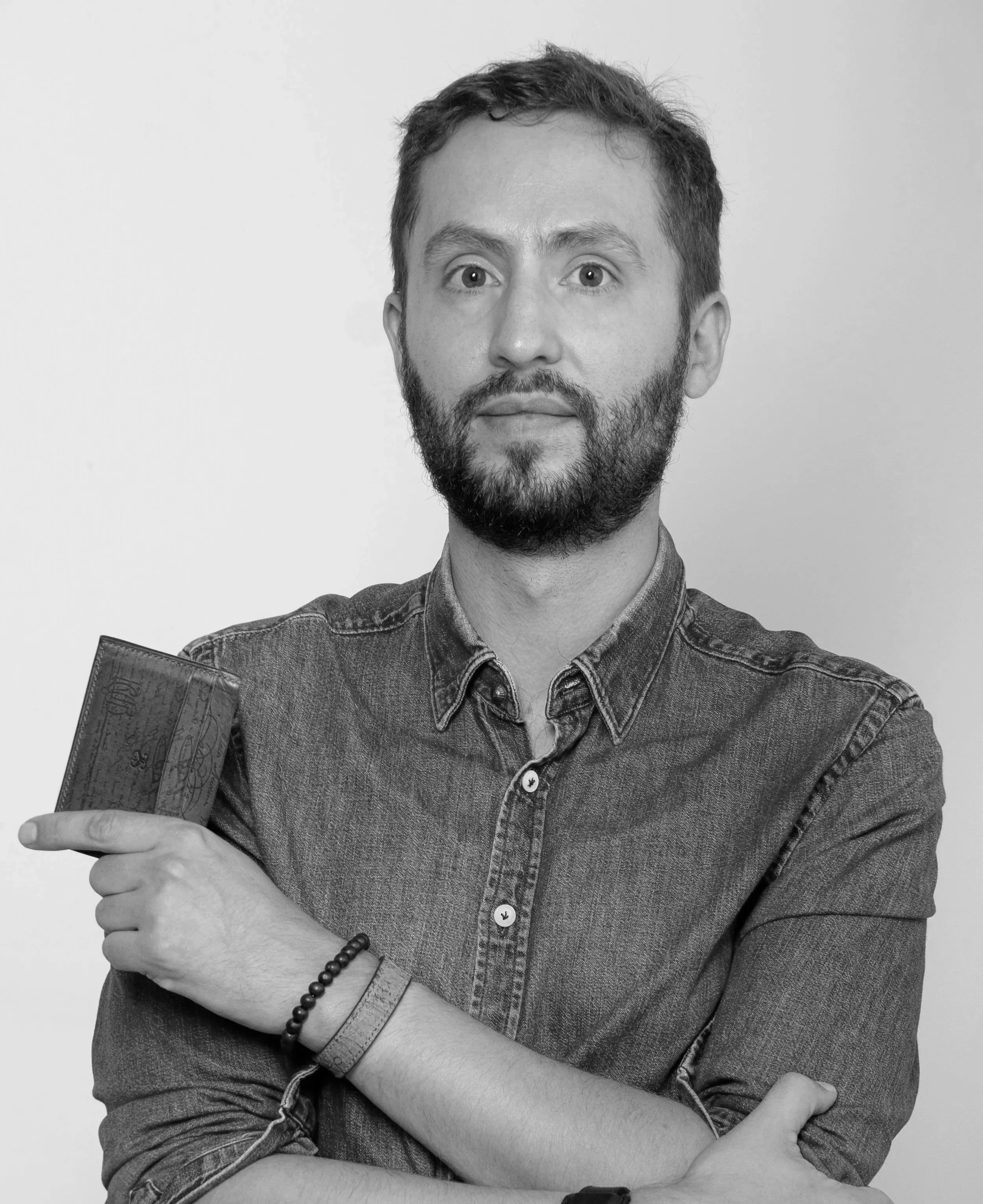 Homme avec barbe, portant une chemise en denim, tenant un portefeuille dans sa main gauche, portant des bracelets et un collier, posé contre un fond blanc.