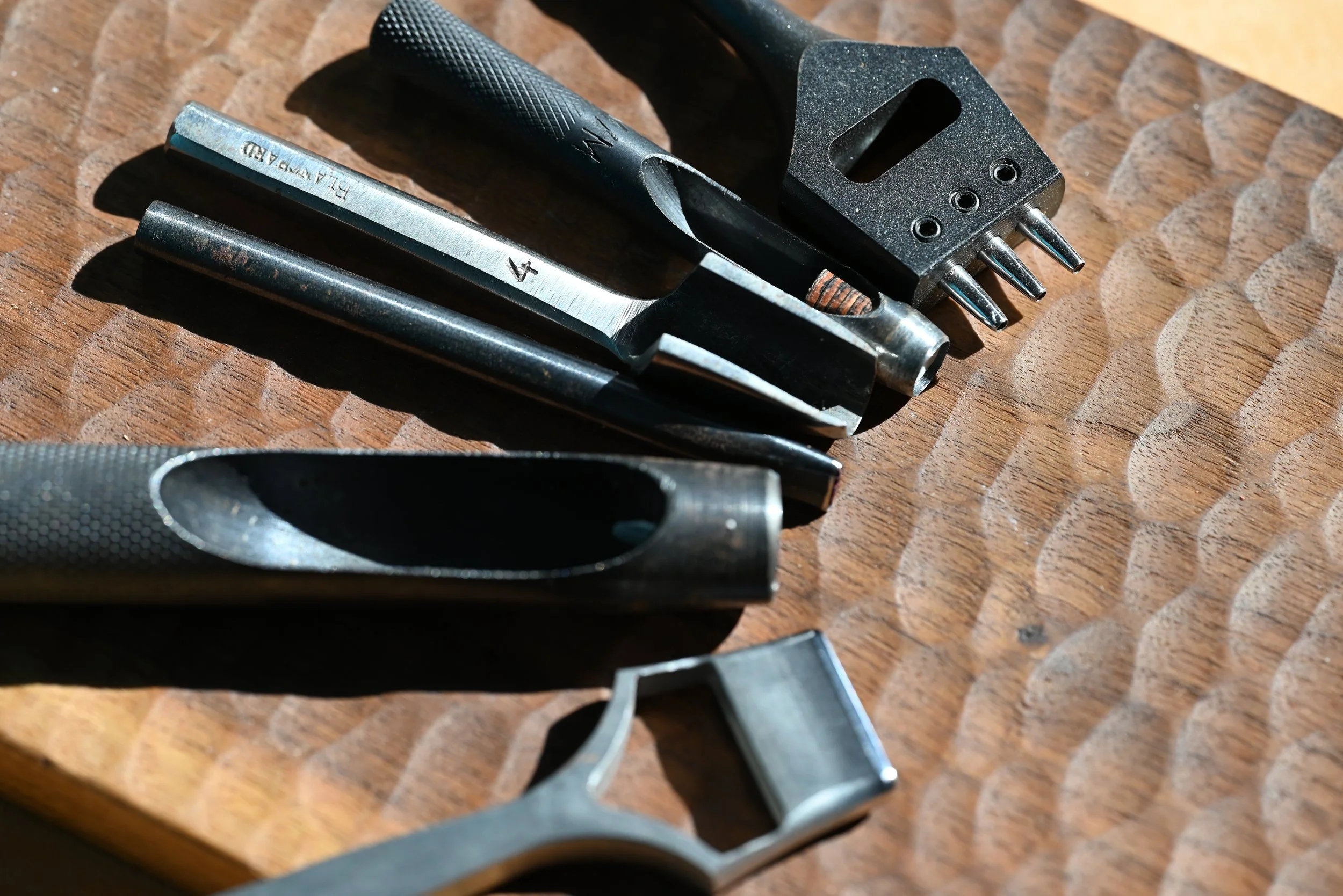 Une variété d'outils de couture ou de bricolage en métal, posés sur une surface en bois, incluant un couteau, une pince, un tournevis et un outil à quatre pointes.
