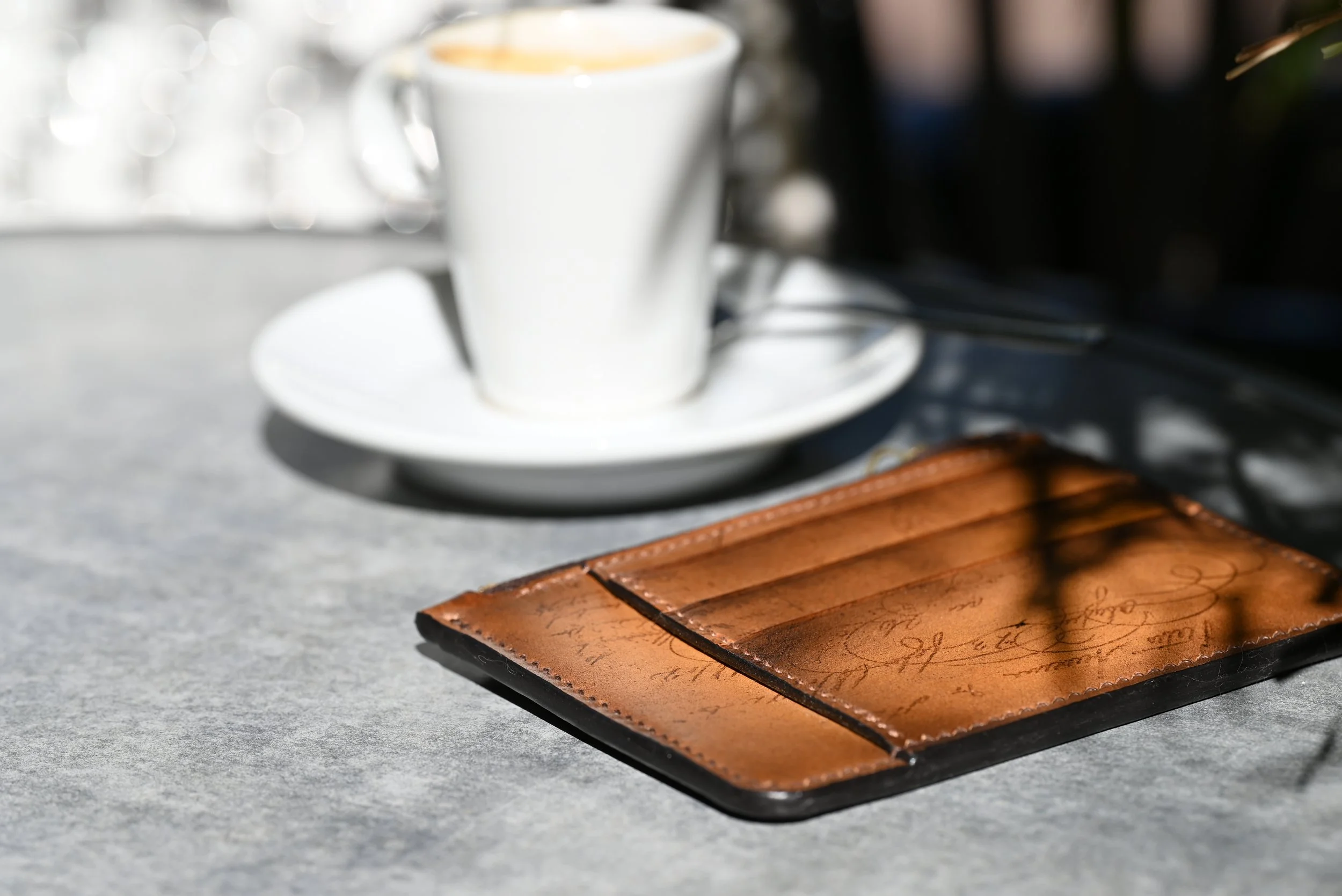Une tasse de café blanc sur une soucoupe blanche avec une cuillère à côté, un portefeuille en cuir marron sur une table en béton.