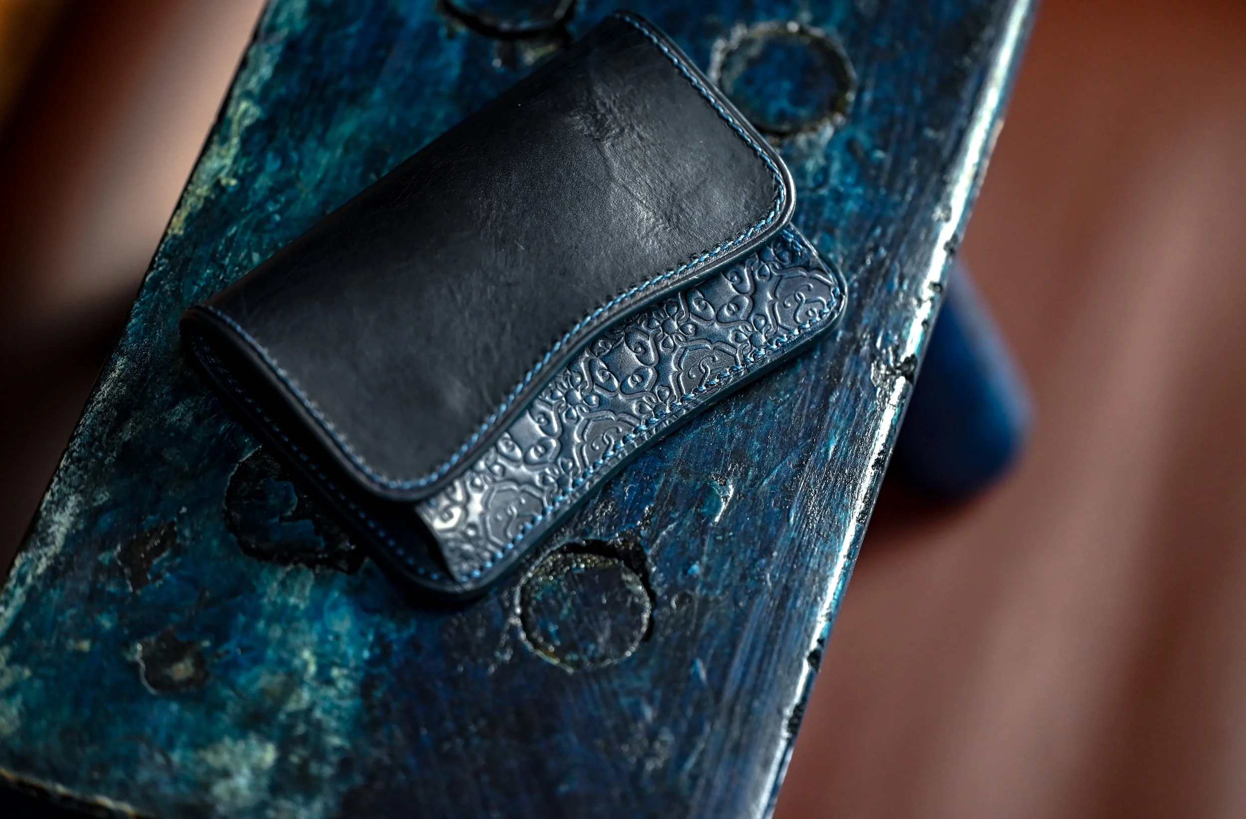 Une petite pochette noire en cuir avec des détails en relief, posée sur un banc en bois peint en bleu délavé.