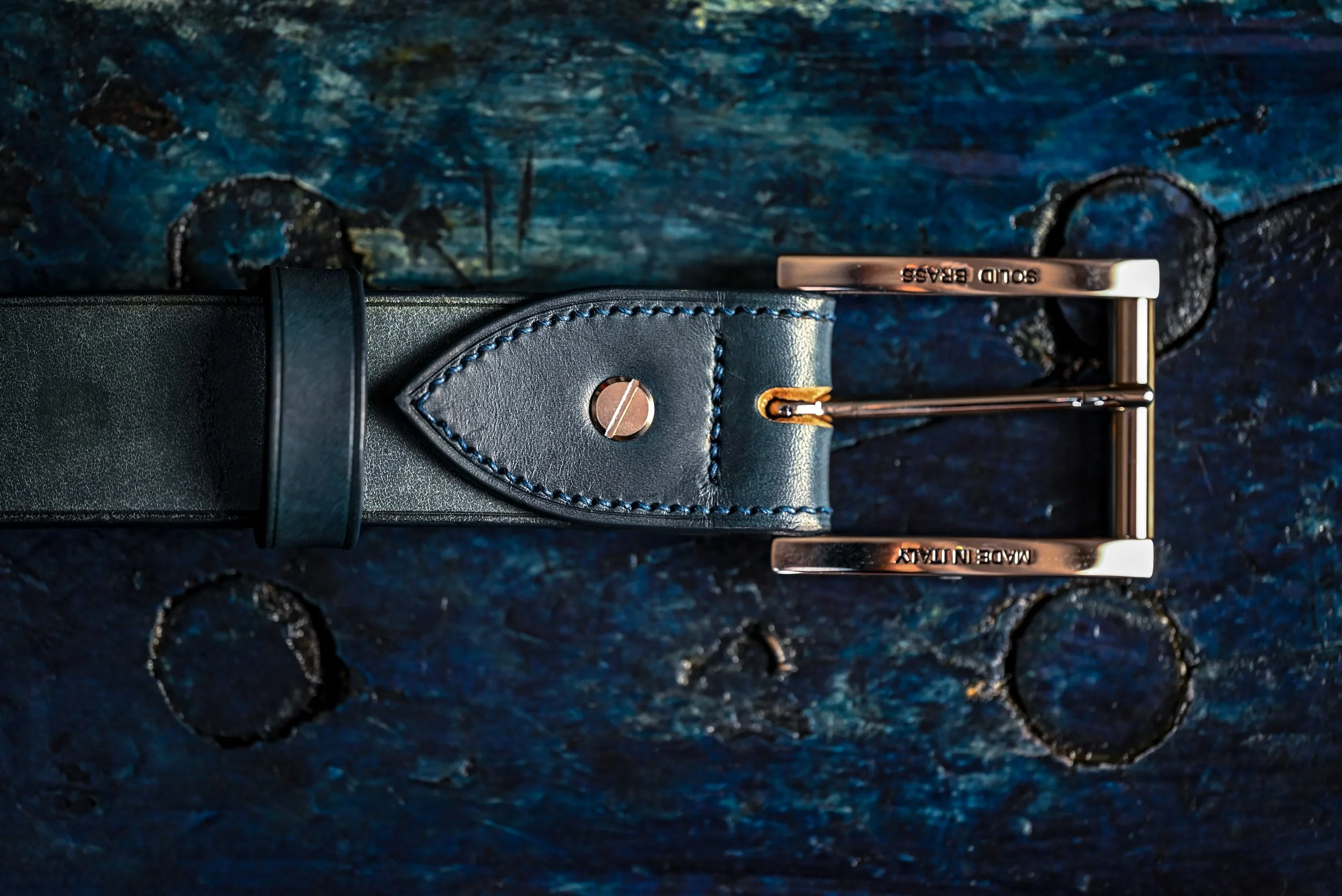 Ceinture en cuir noir avec boucle en métal inscriptée  "MADE IN ITALY" et "SOLD BASS". La ceinture repose sur une surface en bois peint en bleu avec des motifs circulaires.