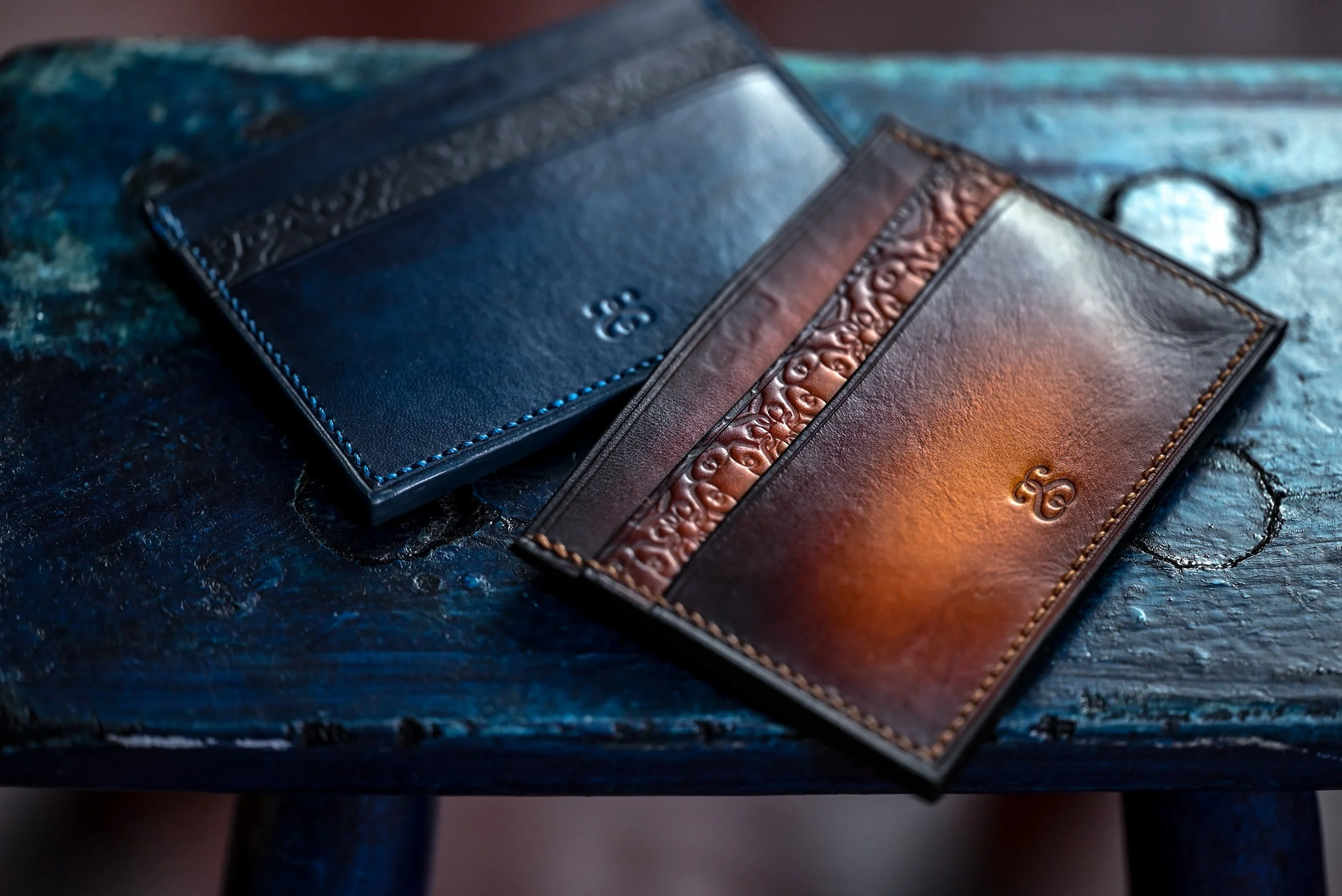 Deux portefeuilles en cuir, l'un bleu avec des coutures bleues et l'autre bicolore brun et noir avec des détails en relief, posés sur une surface en bois foncé.