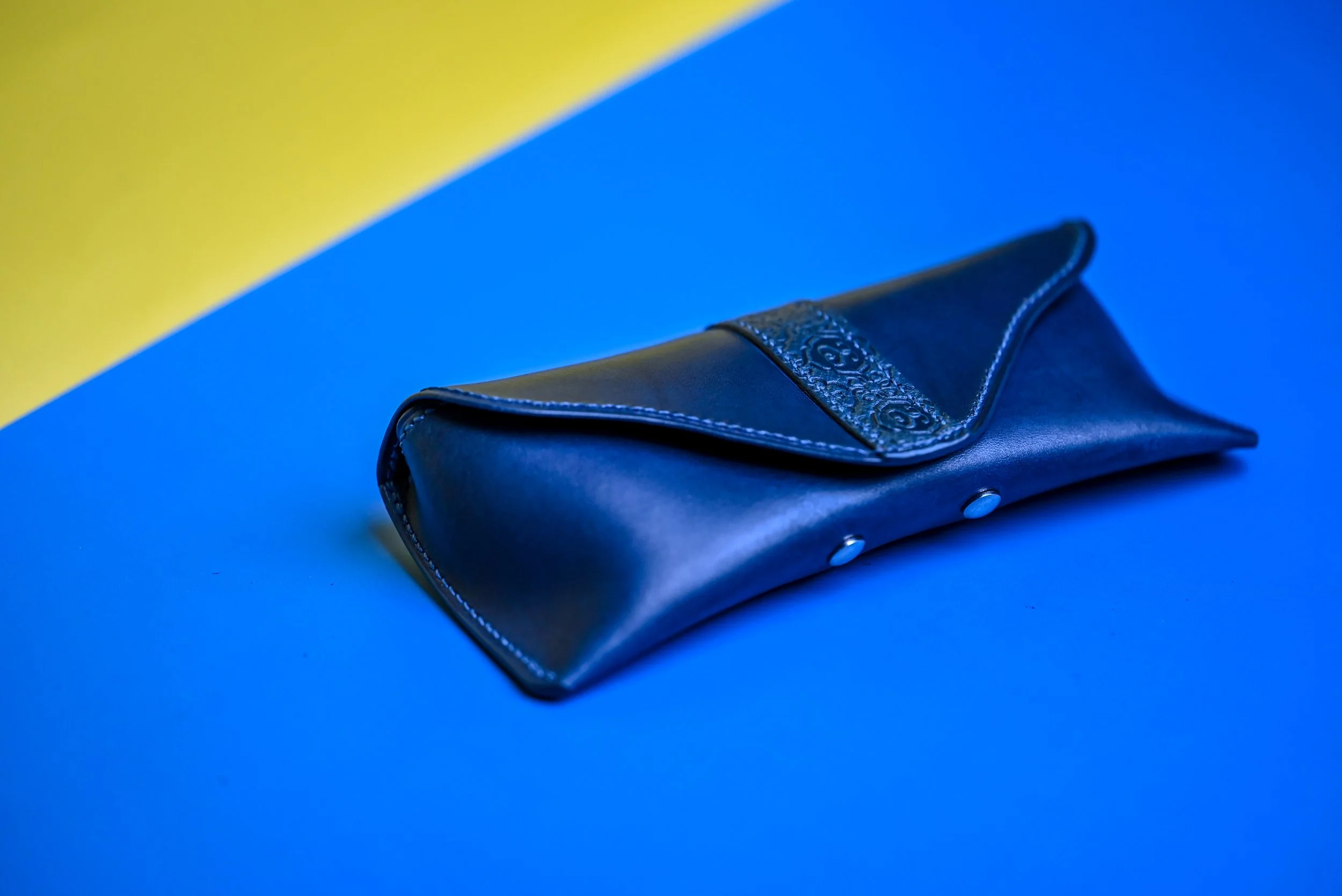 Une pochette noire en cuir avec un rabat décoré, posée sur une surface bleue avec un fond à moitié jaune.