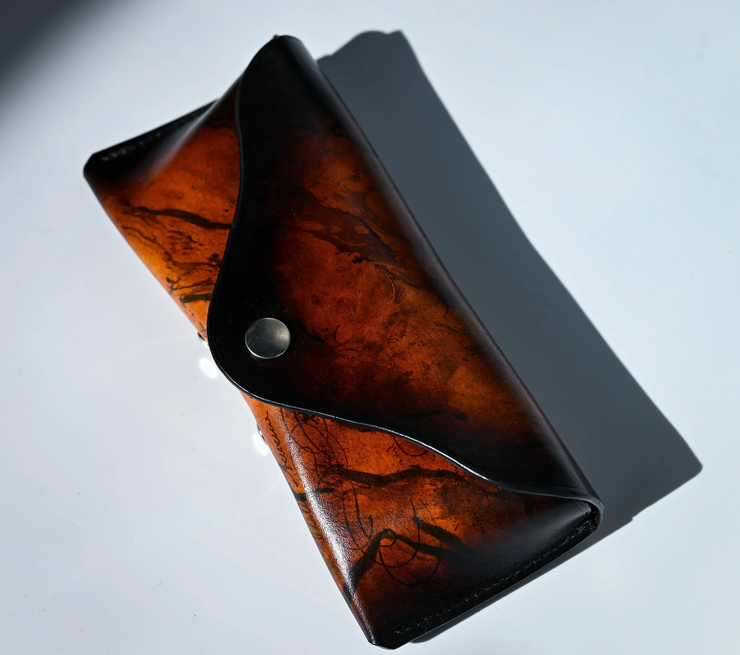 Étui pour lunettes en cuir orange et noir avec un design artistique et une bride à bouton-pression.