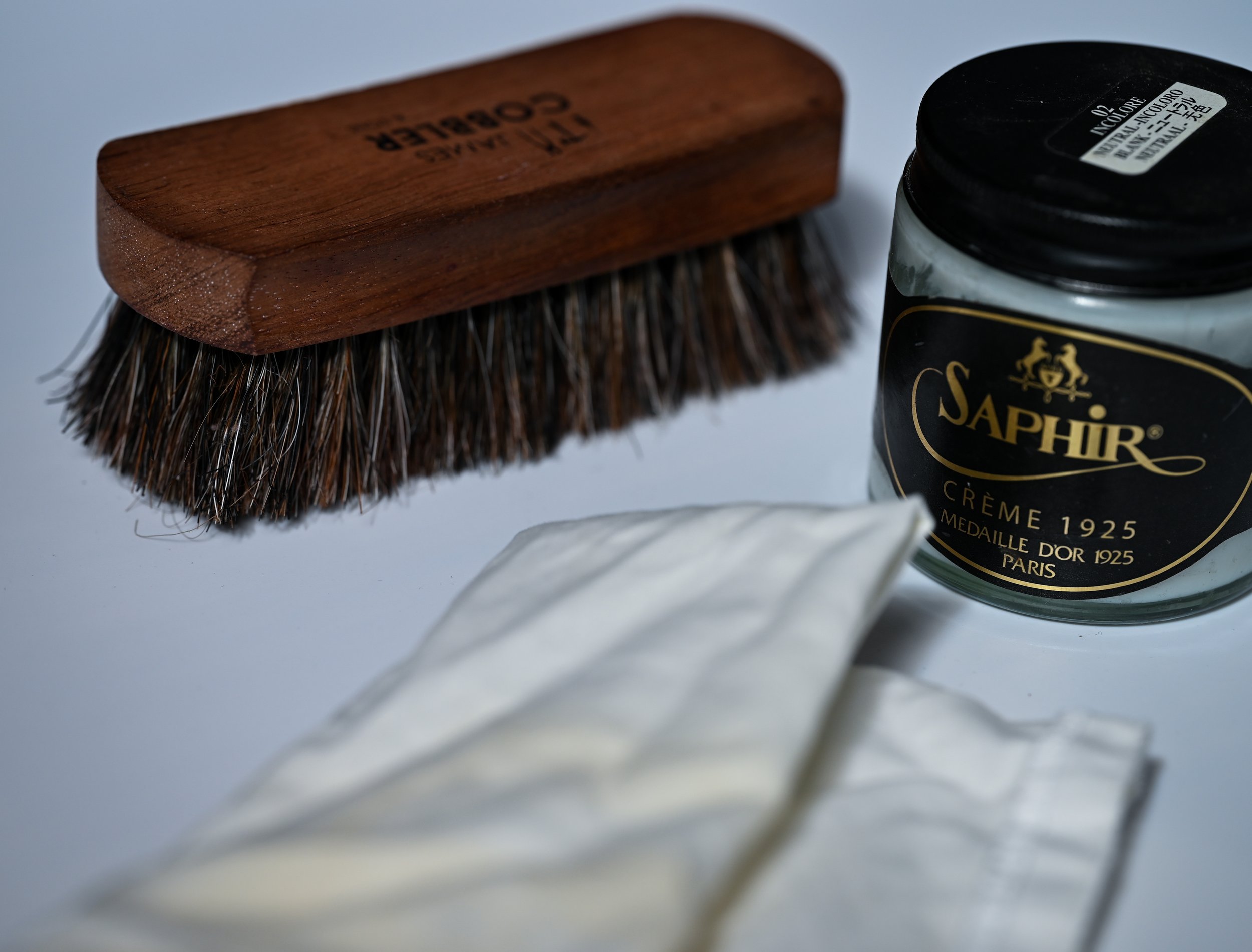 Une brosse à chaussures en bois, un pot de crème Saphir Médaille d'Or, et une bouse en tissu blanc.