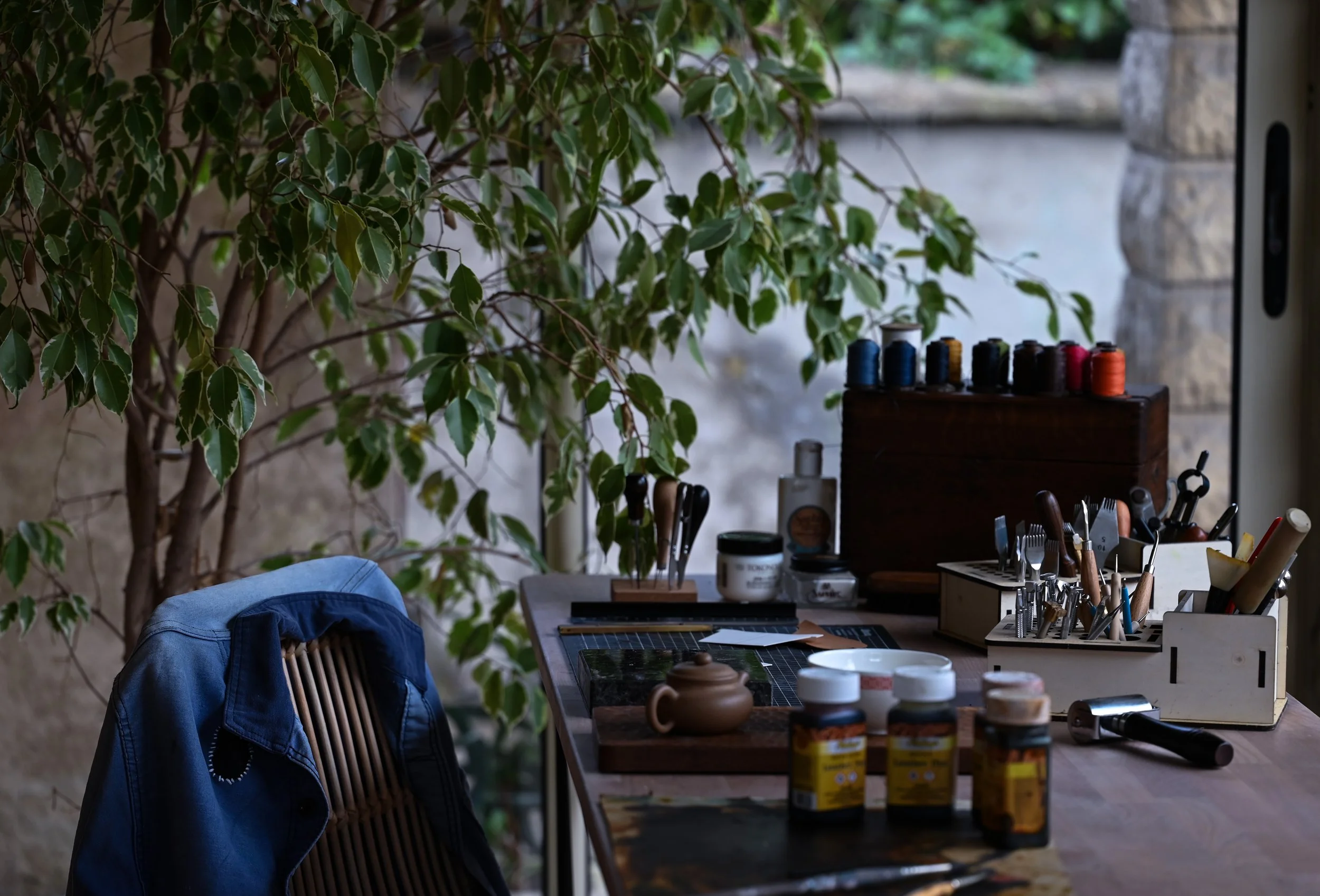 Atelier de travail de Darven avec une table remplie d'outils de maroquinerie, une chaise avec une veste de travail et une plante au fond de la pièce