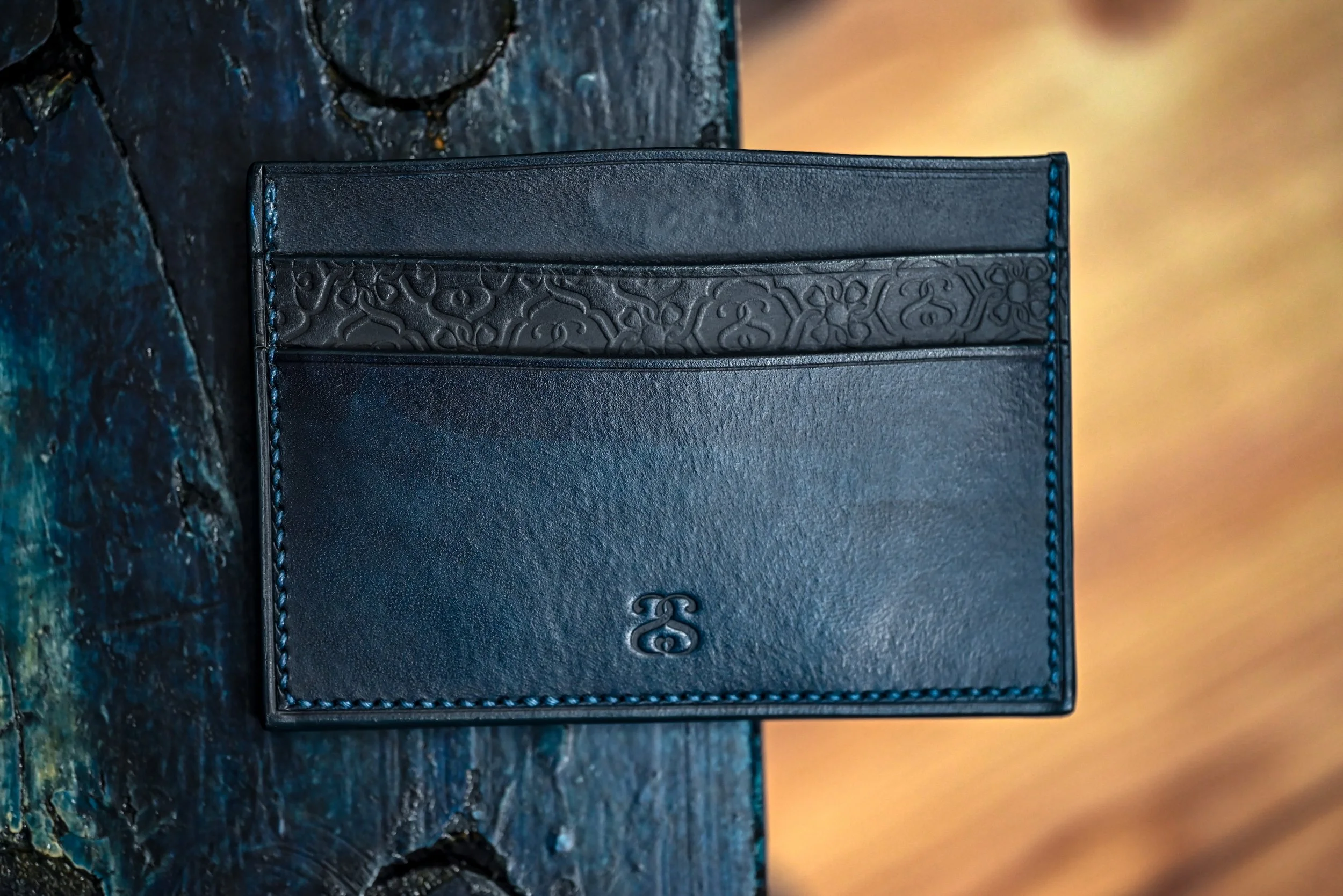 Une portefeuille en cuir noir avec une broderie sophistiquée dans la partie supérieure et un logo discret en bas au centre, posé sur une surface en bois foncé.