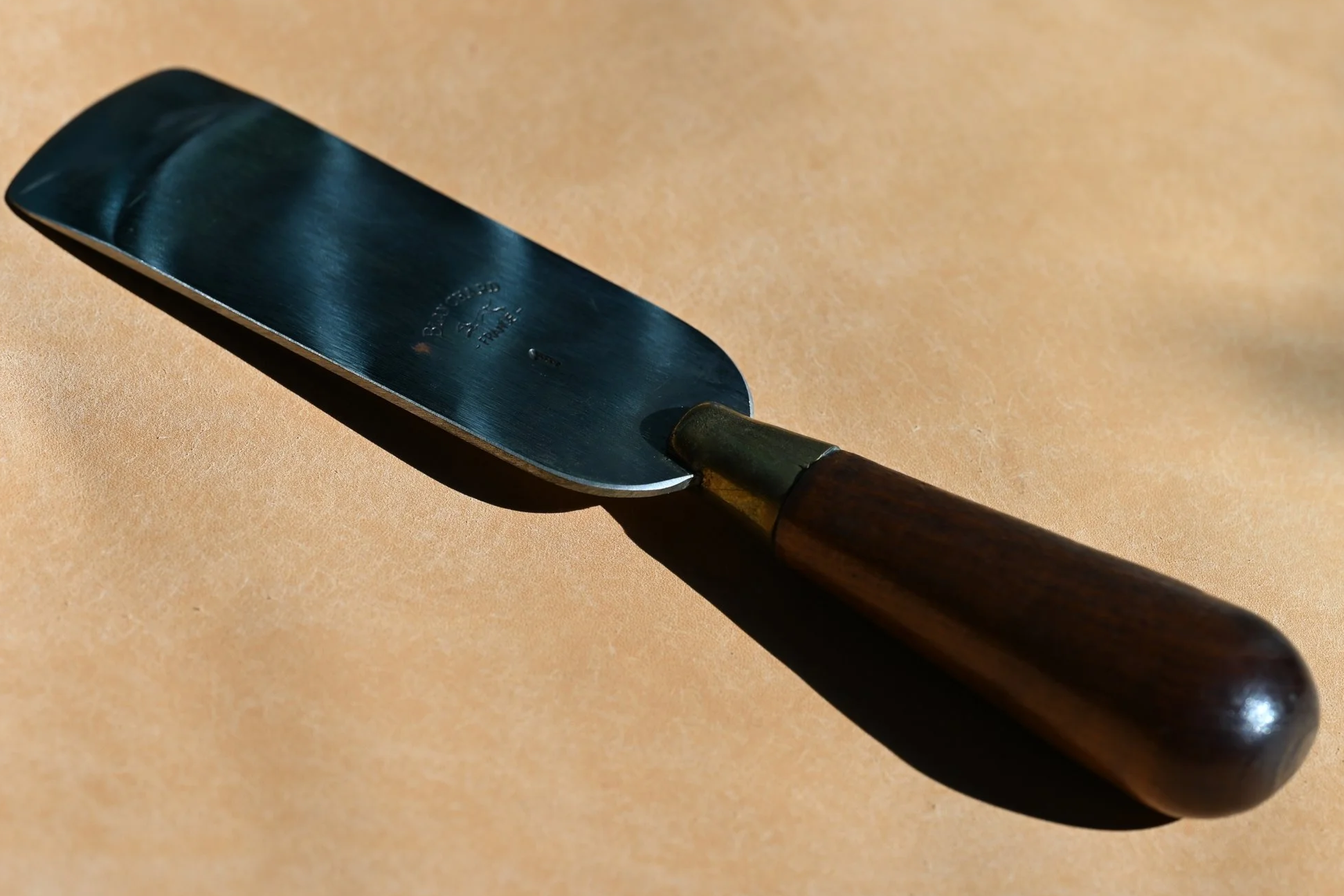 Lame à parer le cuir en métal avec un manche en bois, posée sur une surface en cuir beige