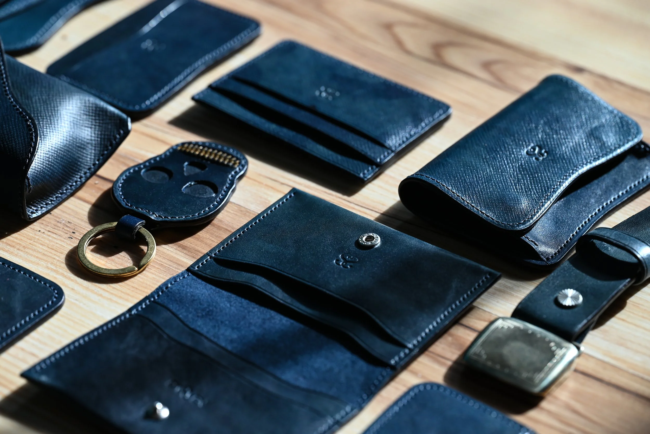 Collection d'articles en cuir noir, comprenant une clé, plusieurs portefeuilles, une pochette, et une montre, étalés sur une surface en bois.
