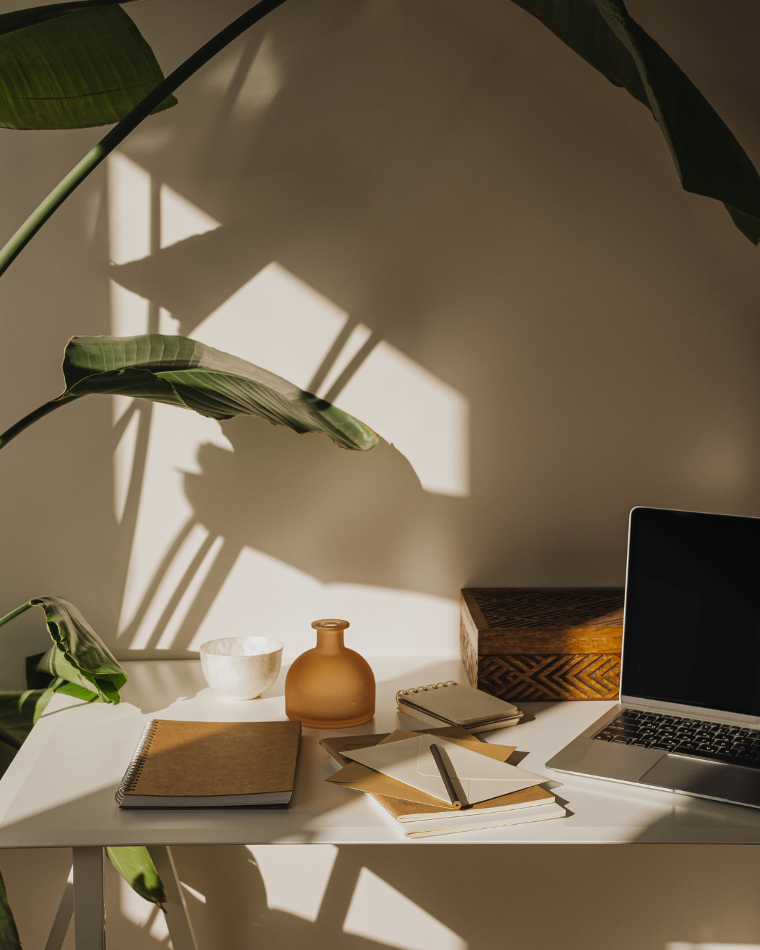 A white desk with a closed laptop, a small notebook, a pen, a decorative glass bottle, and a wooden box. Sunlight creates shadows of large green leaves on the wall behind the desk.