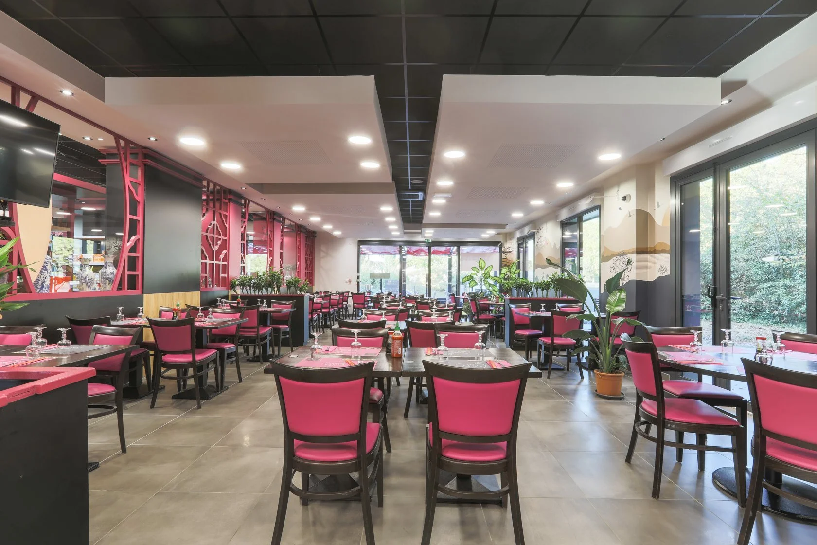 Salle à manger d'un restaurant moderne avec des tables et chaises rose, grandes fenêtres laissant entrer la lumière naturelle, et des plantes d'intérieur.