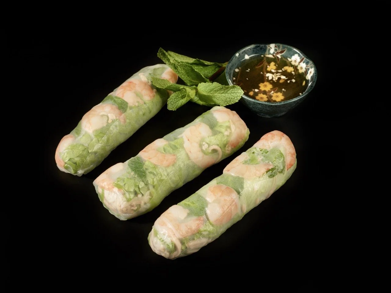 Trois rouleaux de printemps avec une sauce en bol, des feuilles de menthe et de la coriandre sur fond noir.