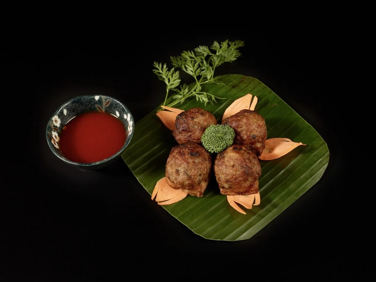 Quatre boulettes de viande frites servies sur une feuille de bananier avec une branche de coriandre, une brocoli, des pétales de fleurs en garniture et une petite tasse de ketchup ou sauce tomate à côté.