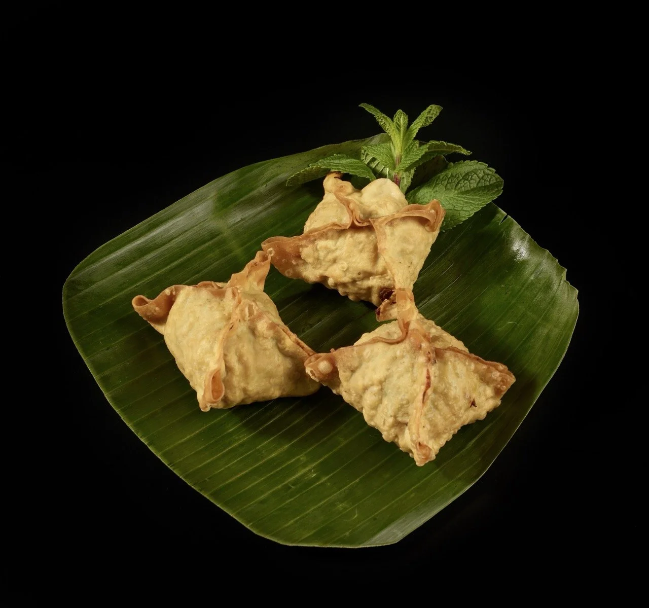 Quatre samoussas frits disposés sur une feuille de bananier avec des feuilles de menthe verte au fond, sur un fond noir