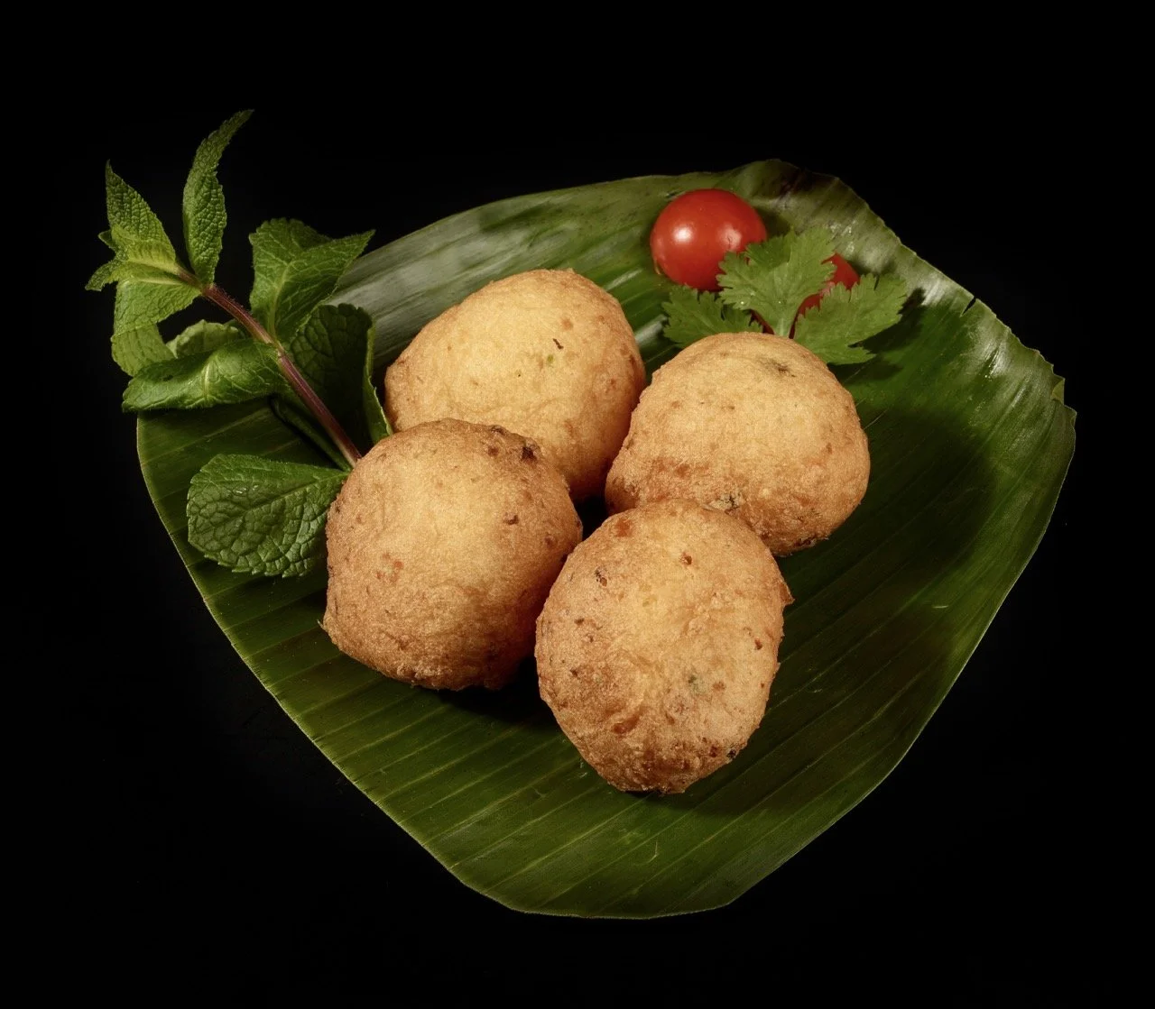 Quatre croquettes dorées sur une feuille de bananier, accompagnées de feuilles de menthe, une tomate cerise, un brin de coriandre et une branche de menthe verte contre un fond noir.