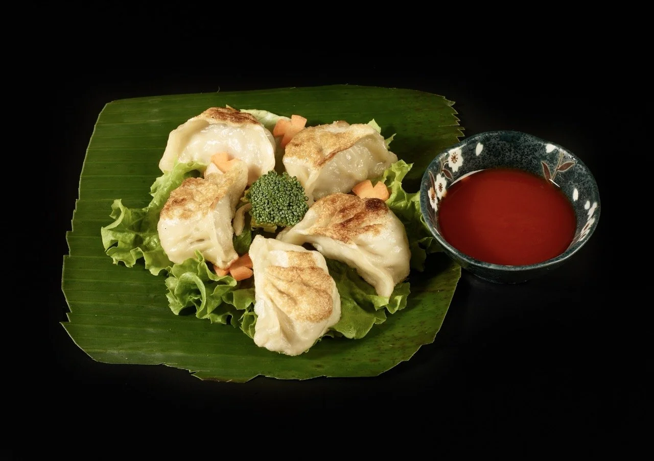 Six dumplings frits servis sur une feuille de bananier avec des légumes verts et une petite tasse de sauce rouge à côté.