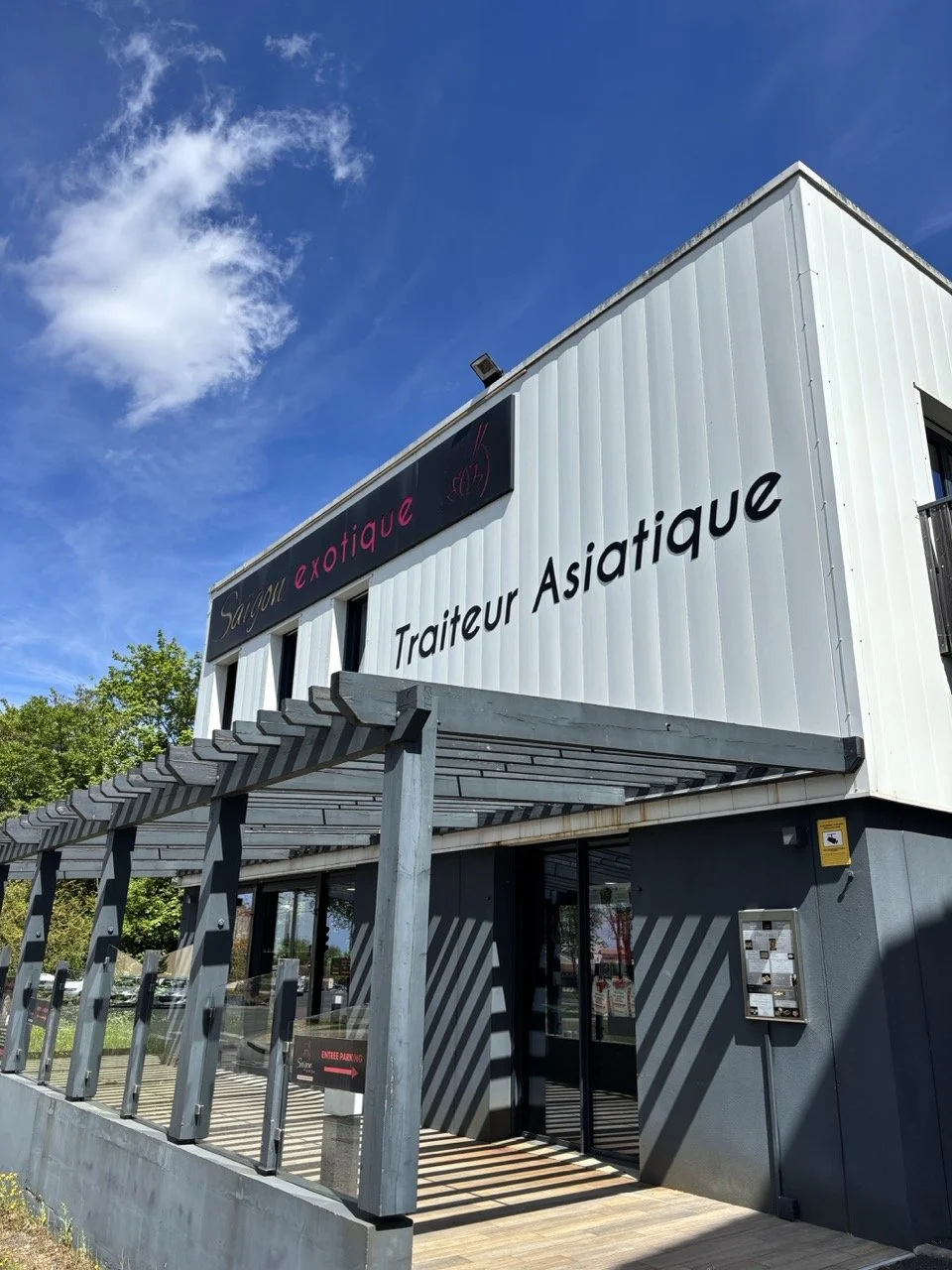 Bâtiment moderne avec une enseigne indiquant 'Salon exotique' et 'Traiteur Asiatique', entrée sous une structure en bois avec une porte en verre et un menu affiché.