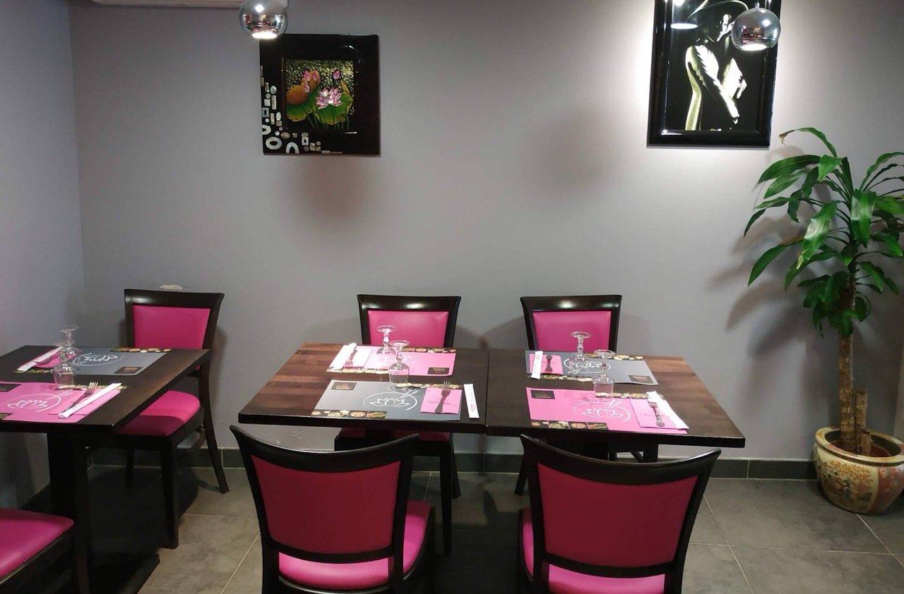 Salle à manger d'un restaurant avec six chaises en bois et coussins roses, trois tables avec menus, fourchettes, couteaux, verres à vin, et assiettes, une grande plante en pot dans le coin, deux tableaux modernes encadrés sur le mur, deux lampes suspendues en métal.