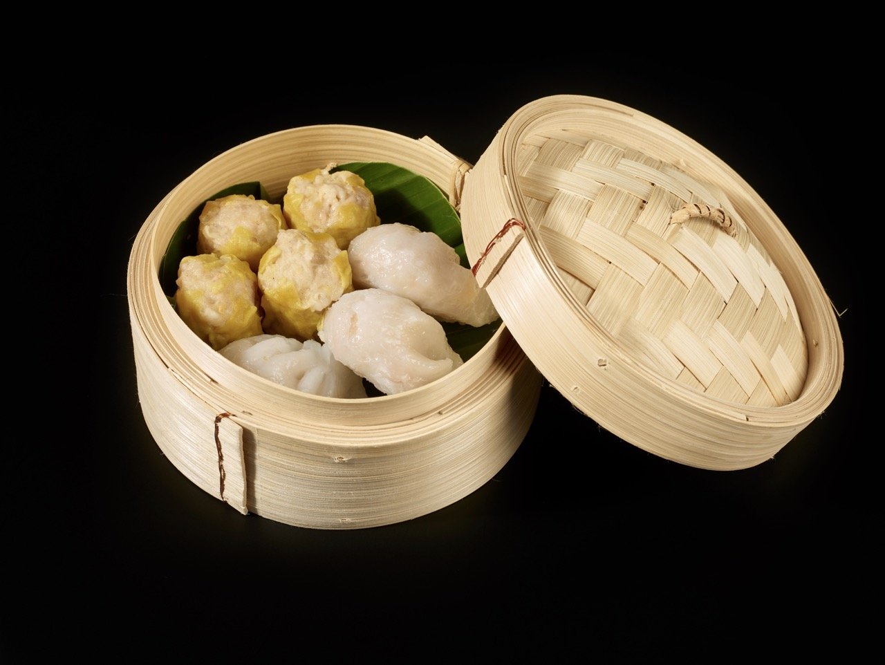 Dim sum dans un panier en bambou avec le couvercle posé à côté, comprenant des siu mai et des har gao sur un fond noir.