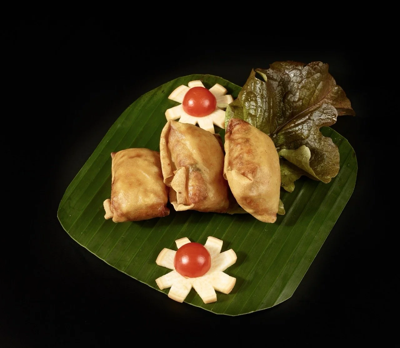 Trios de nems frits sur une feuille de bananier avec de la salade verte, accompagnés de décorations de légumes en forme de fleurs avec des tomates cerises.