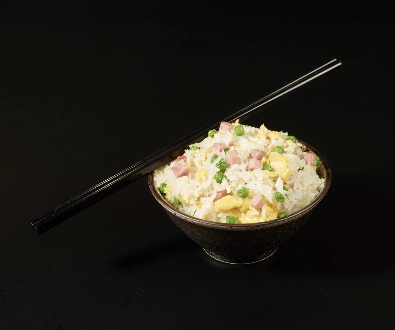 Bol de riz frit avec petits morceaux de jambon, œufs et pois verts, avec des baguettes posées dessus, sur fond noir.