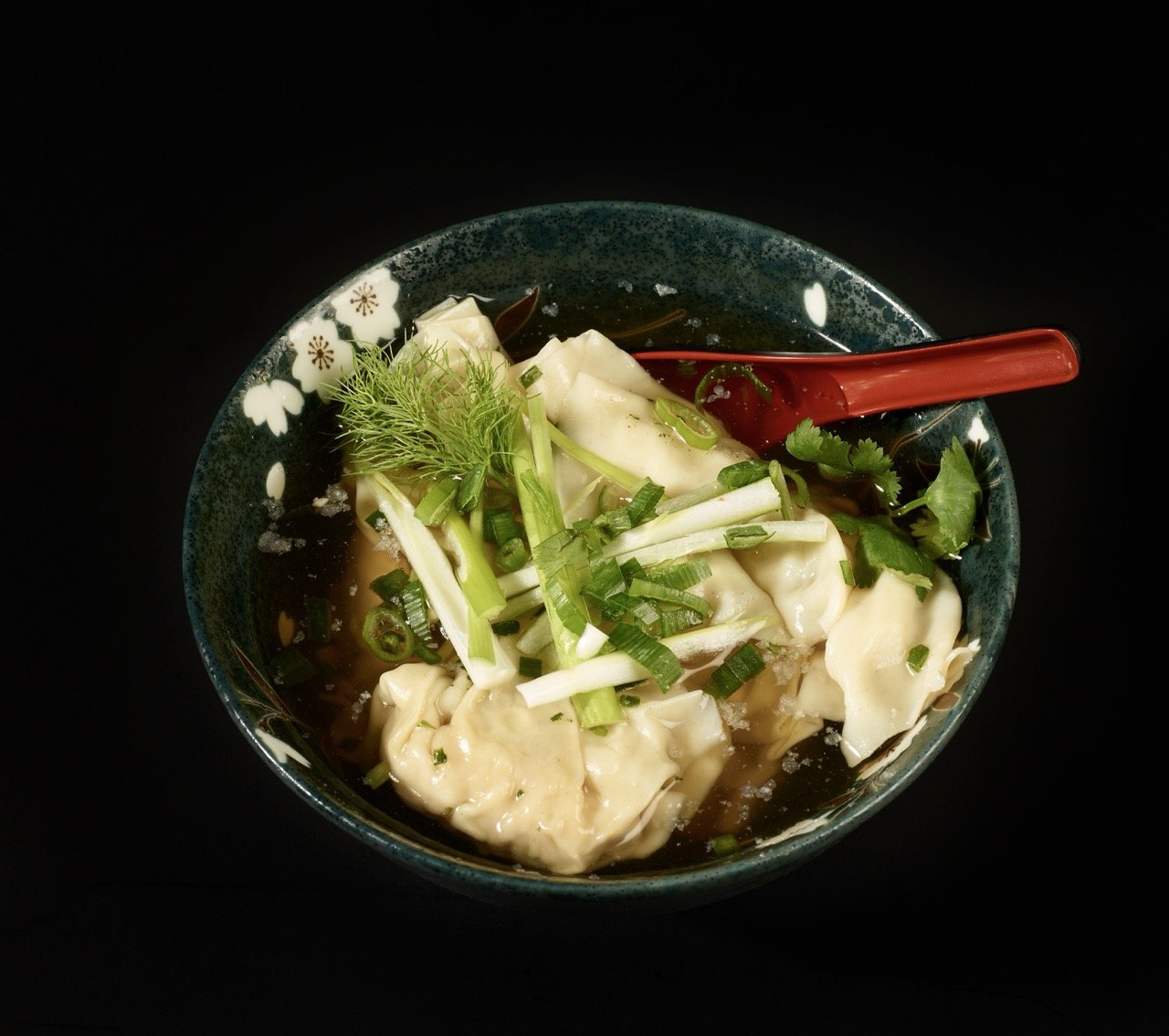 Bol en céramique contenant des dumplings chinois garnis de ciboulette, coriandre, et herbes, avec sauce soja, sur un fond noir.