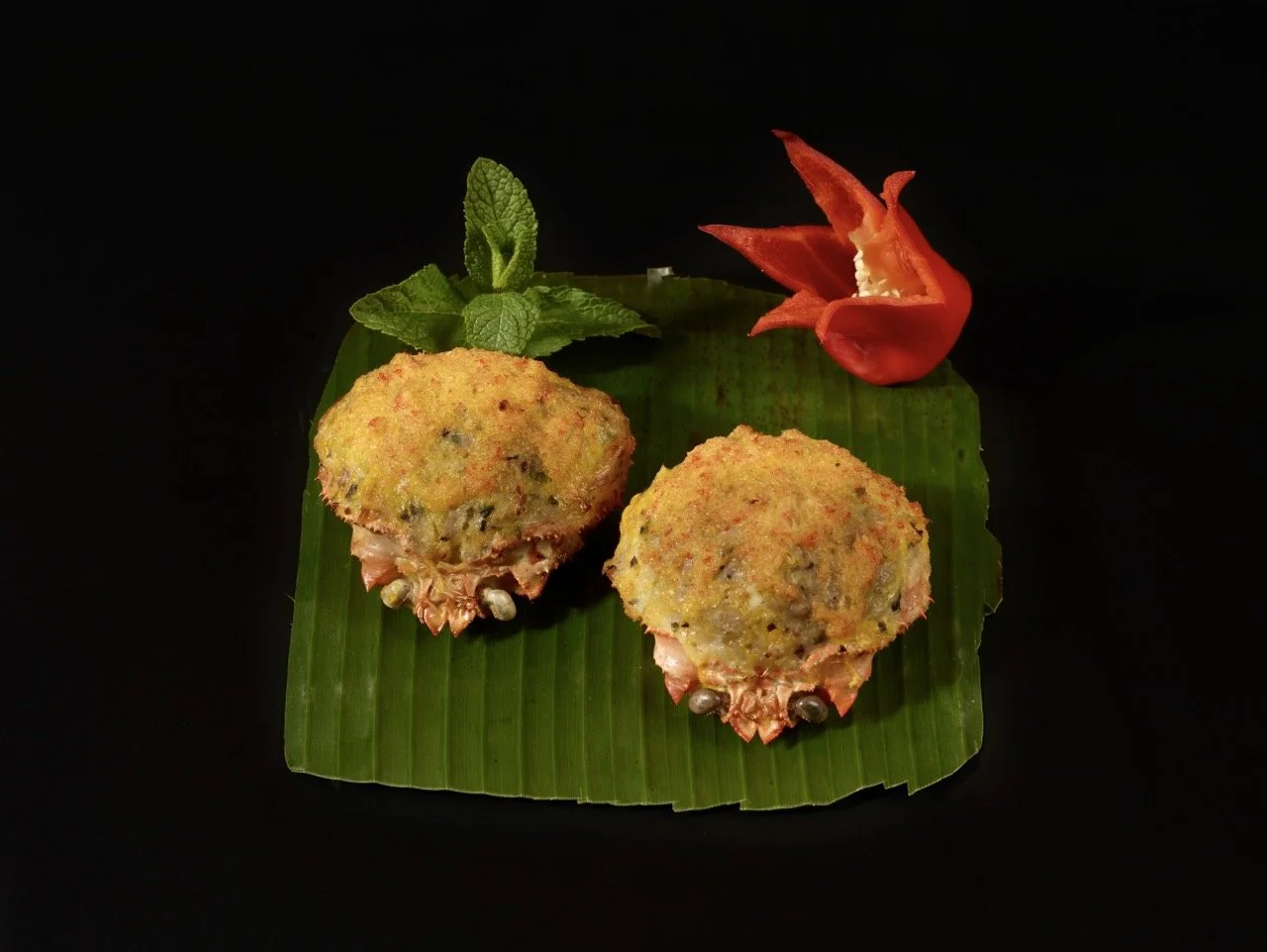 Deux croquettes de crabe sur une feuille de bananier avec une branche de menthe, une fleur rouge et un fond noir.